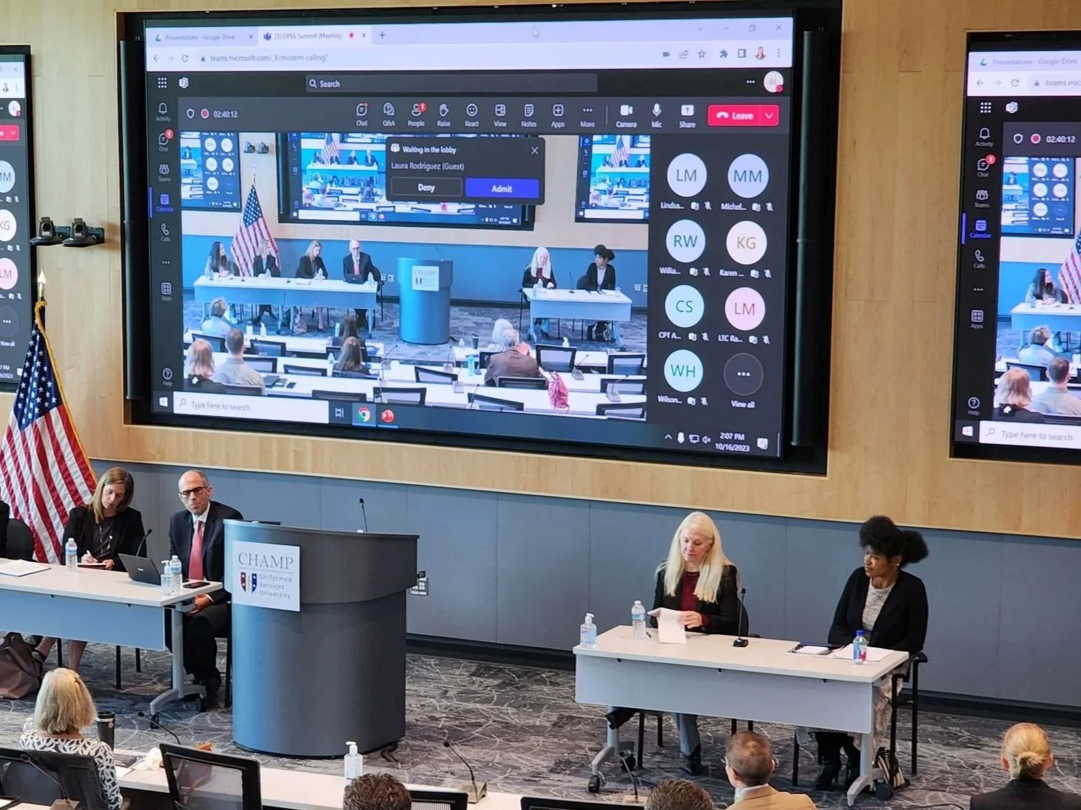 A conference room with a large screen projector displaying a virtual meeting with multiple participants. Several people are seated at tables, listening and taking notes, some using laptops or talets.