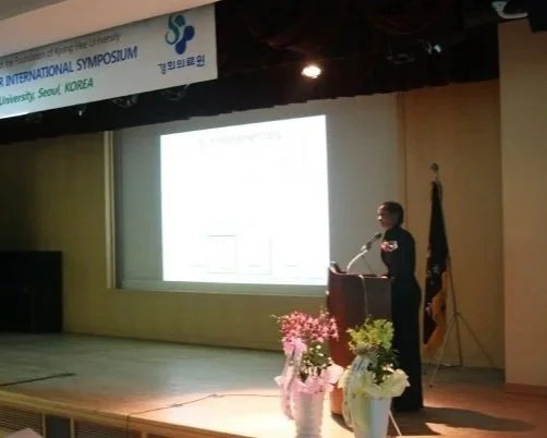 A person giving a presentation at an international symposium in Seoul, Korea. They are standing at a podium with a microphone, with flowers on the stage and a large screen behind them displaying a presentation slide.