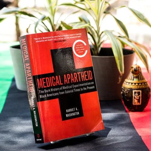 A book titled 'Medical Apartheid' by Harriet A. Washington standing upright on a table with plants and a decorative vase in the background.
