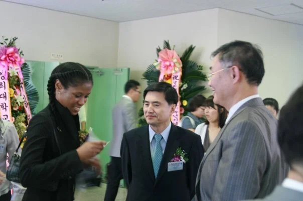 Three people engaged in conversation at a formal event, with floral arrangements and other guests in the background.