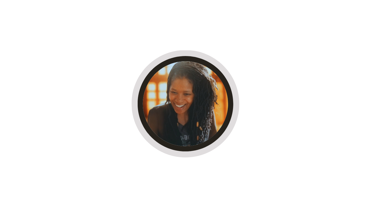 A woman with dark, curly hair styled in locks, smiling and looking down, with warm lighting and blurred background.