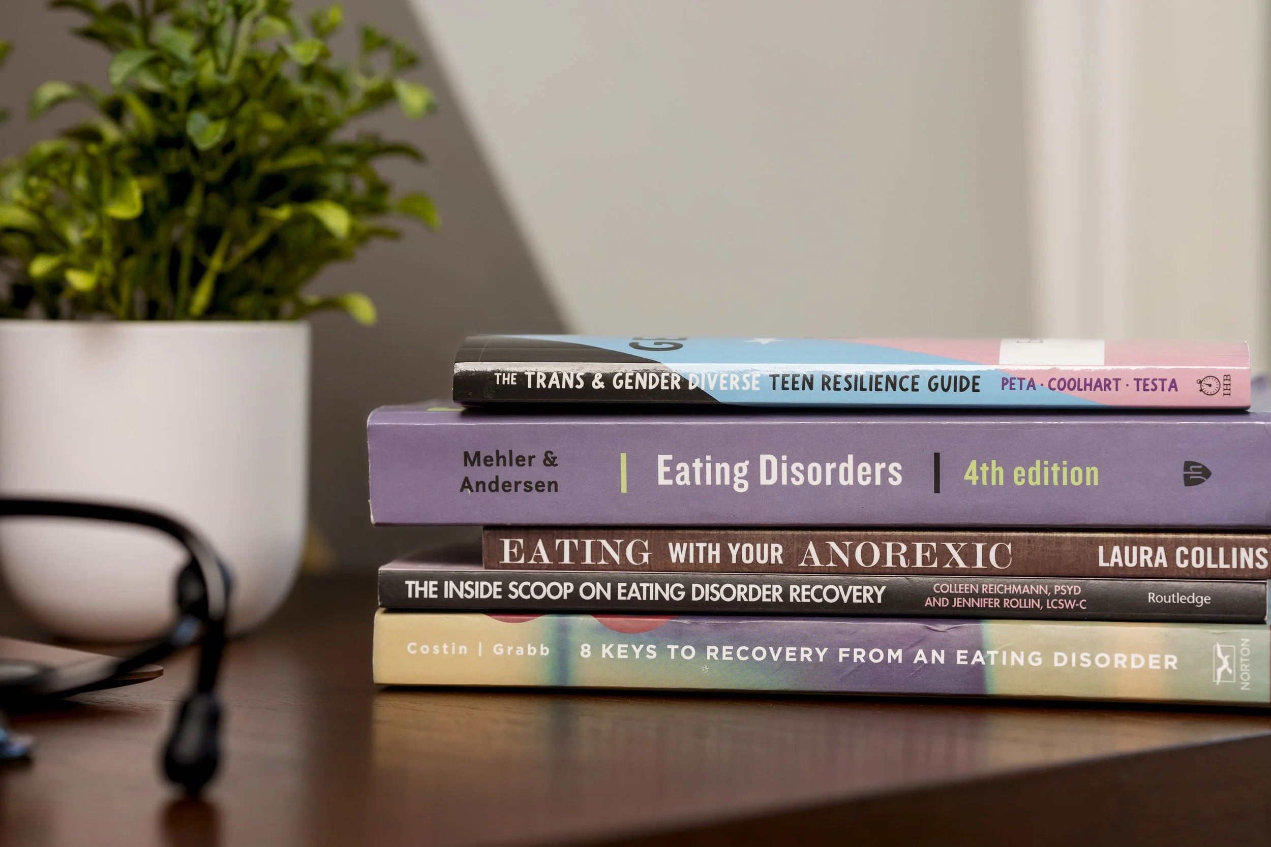 Stack of five books on a wooden surface with a potted green plant in the background.
