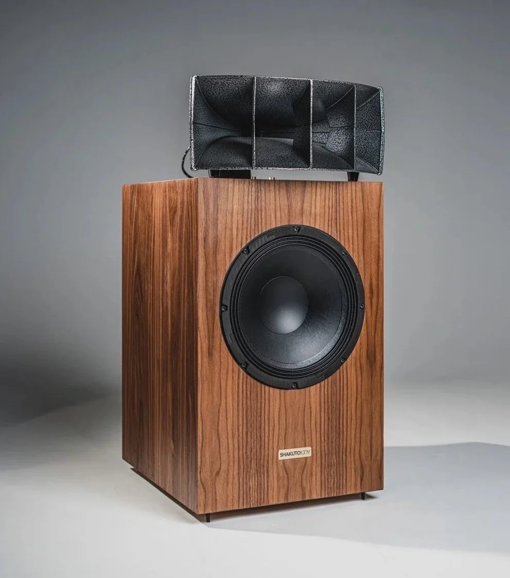 A large wooden speaker with a prominent cone, sitting on a beige carpet in a room with black acoustic panels and a window with curtains.
altec 811b horns 