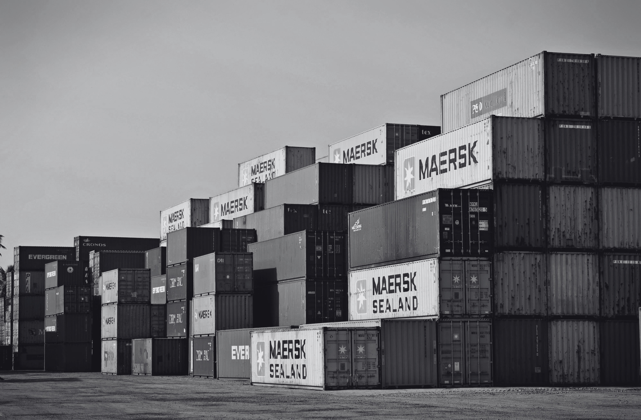 Stacked shipping containers at a port, with logos like Maersk and Evergreen visible.
