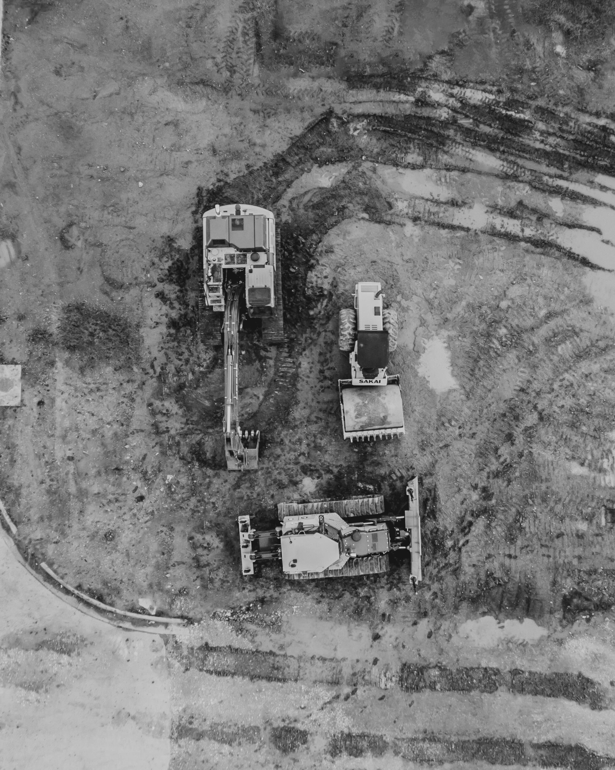 Black and white aerial view of construction vehicles on a dirt site, with two excavators and a road grader.
