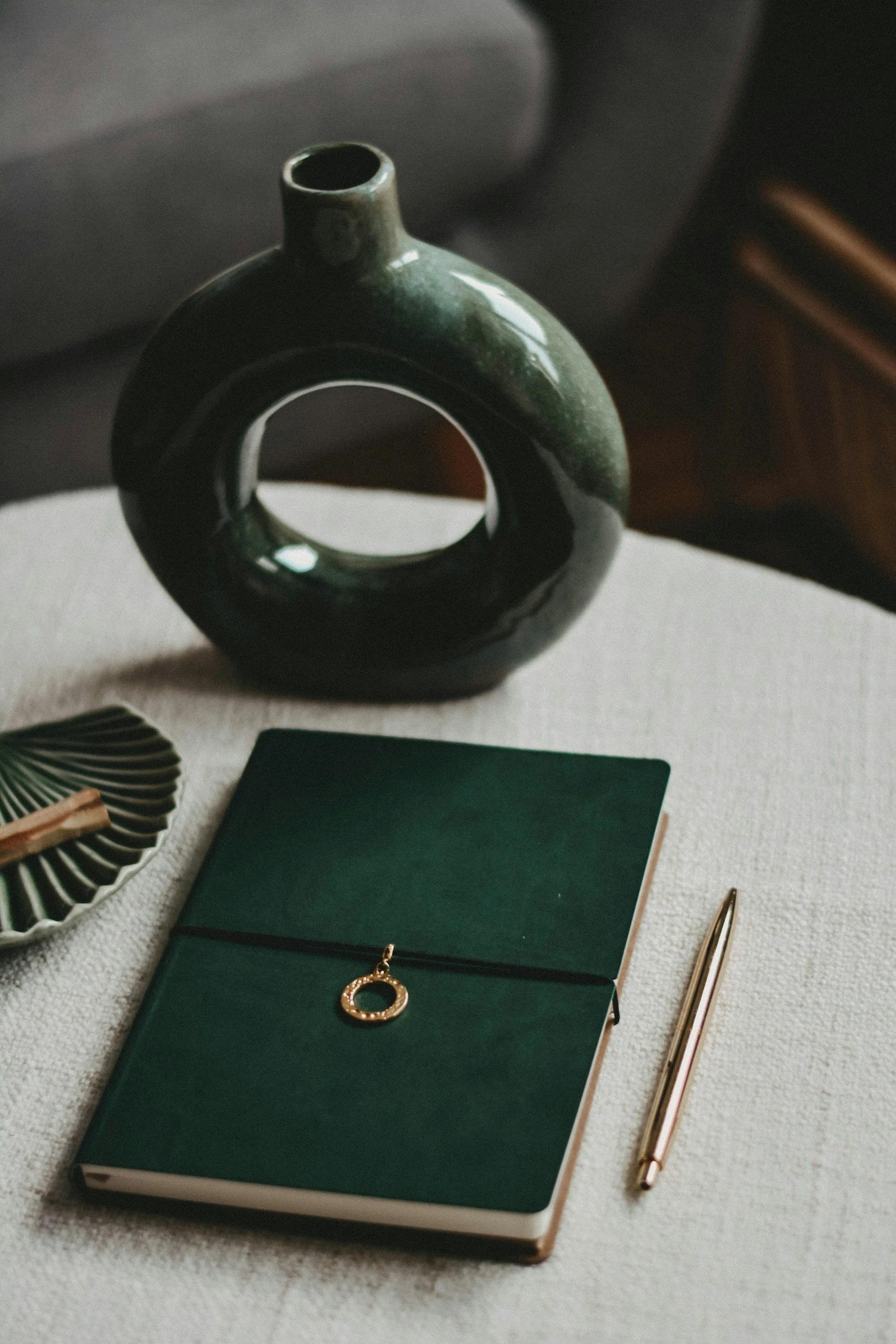 A green notebook with a gold ring-shaped bookmark, a gold pen, a dark green vase with a circular hole, and a small dish with a wooden clothespin and a decorative green leaf, all arranged on a white textured surface.