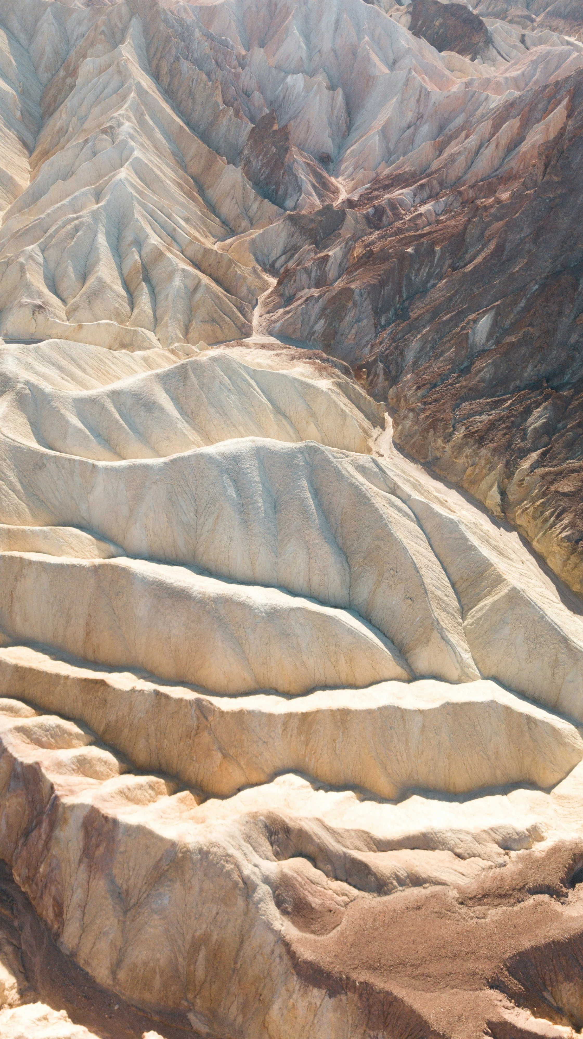 Aerial view of layered, multicolored desert mountains with light tan, white, brown, and reddish hues and deep valleys running through the landscape.