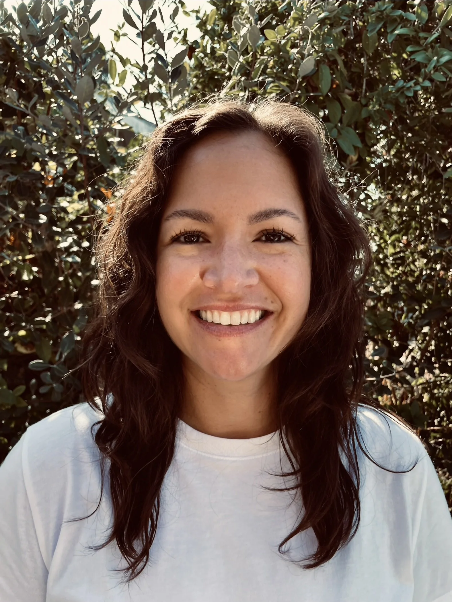 Smiling woman with wavy brown hair standing outdoors in front of leafy bushes, wearing a white T-shirt.
