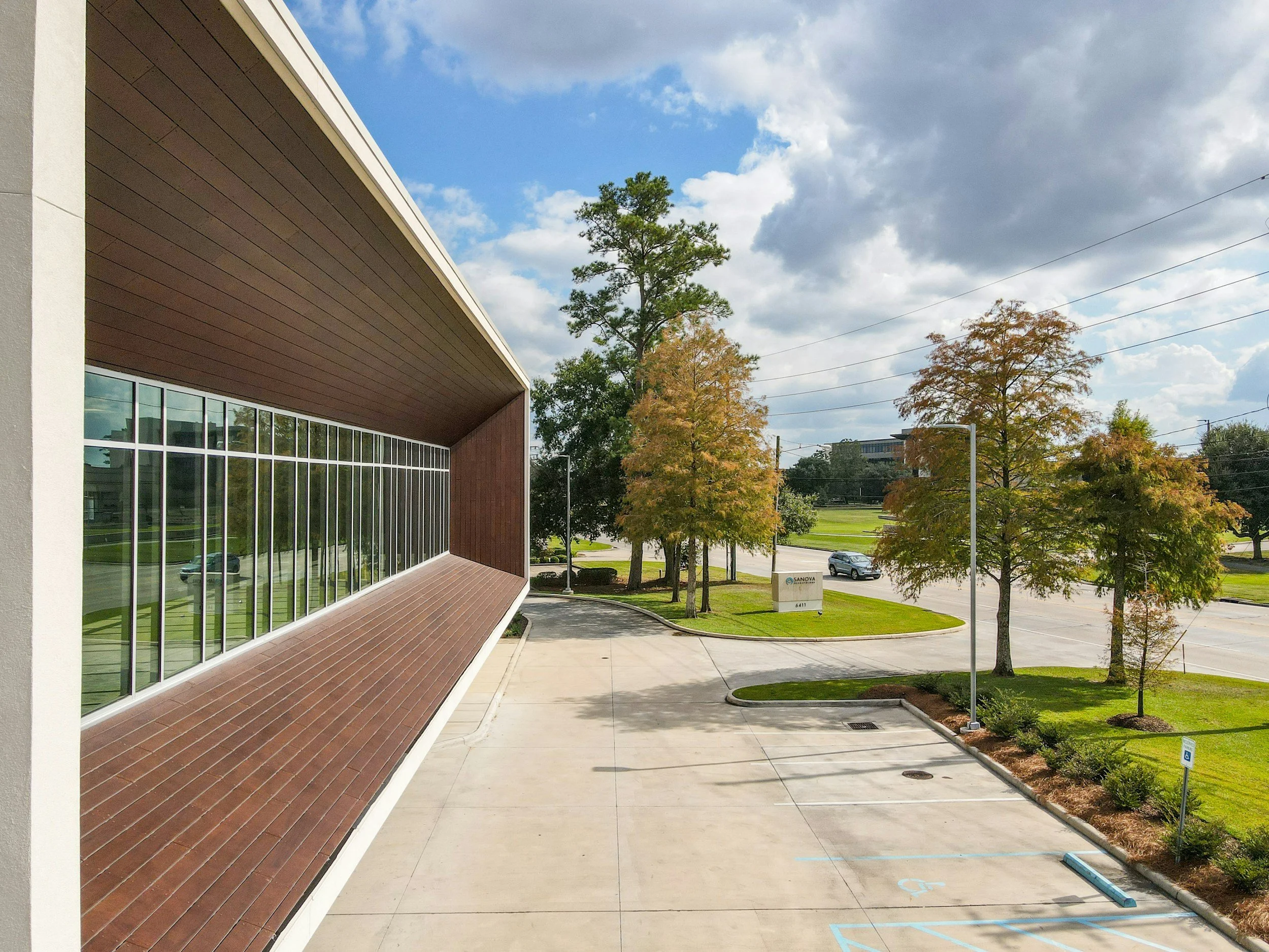 Exterior view of a modern building with large glass windows and a wood-paneled overhang. The area has trees with changing leaves, a sidewalk, and a parking lot with a few cars. The sky is partly cloudy.
