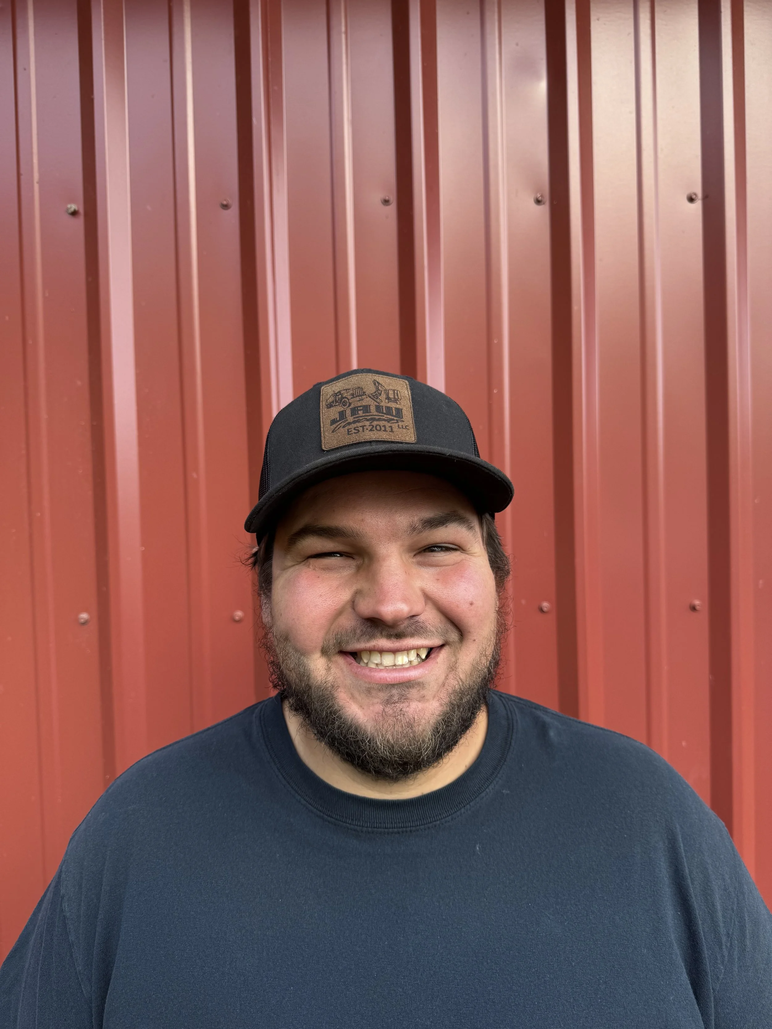 A smiling man with a beard wearing a black cap and a navy blue T-shirt standing in front of a red metal wall.