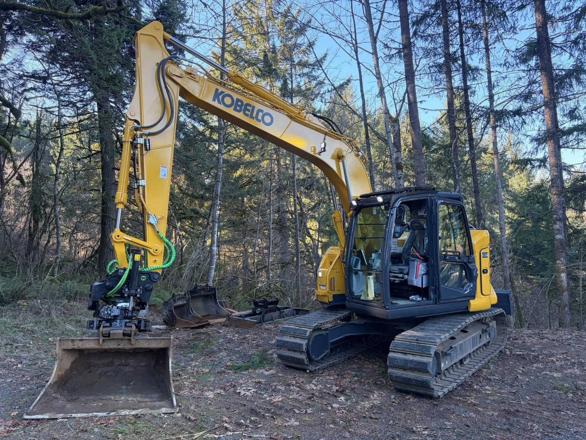A yellow Kobelco excavator with black cabin tracks is parked in a wooded area with trees and clear sky in the background.