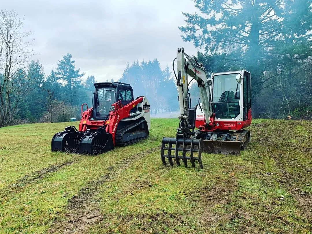 Two small pieces of construction equipment, a skid steer on the left and a mini excavator on the right, parked on a grassy field with a wooded background and cloudy sky.