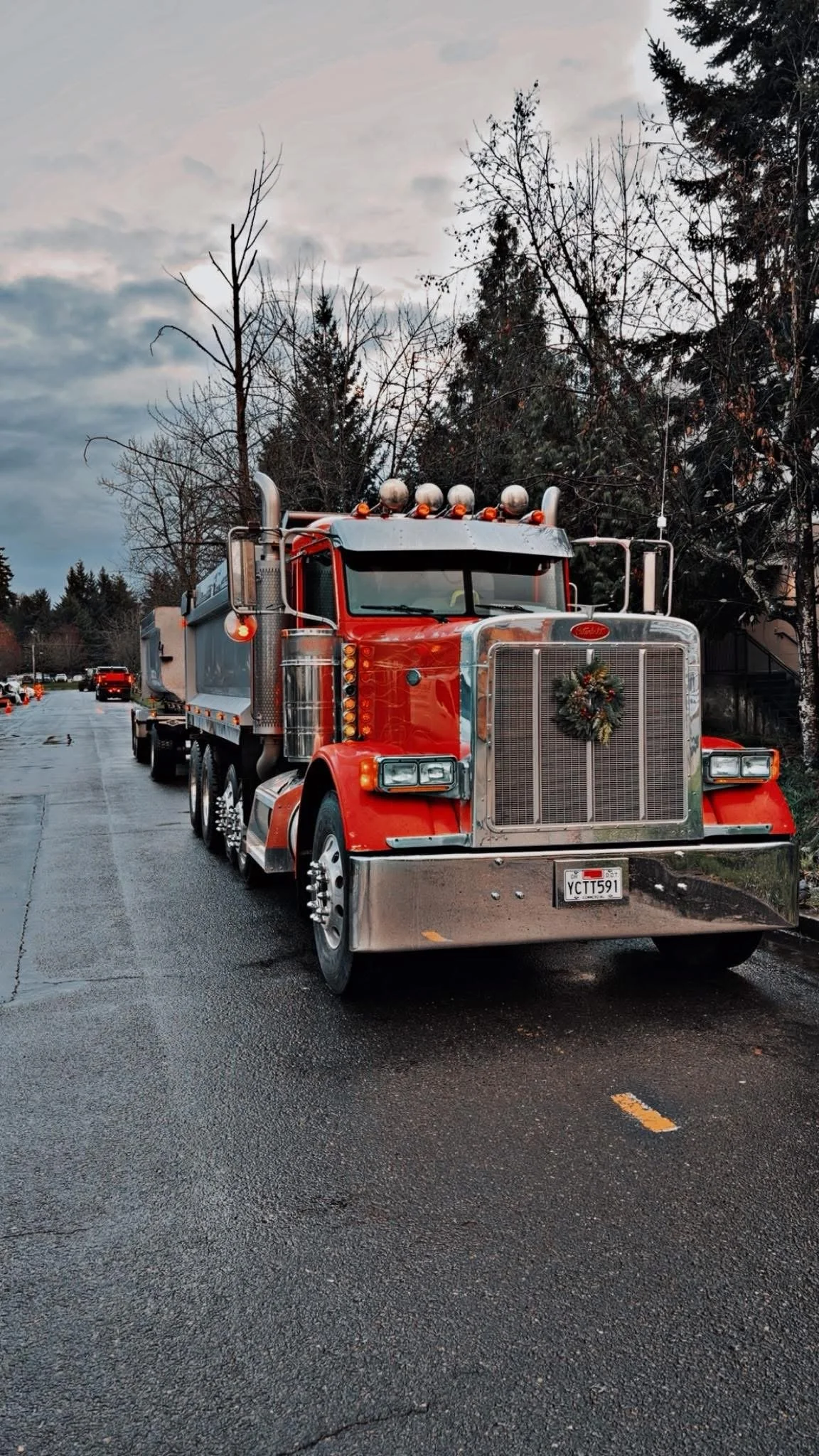 A red fire truck with a holiday wreath decoration on its front grille parked on a wet street during the cloudy evening.