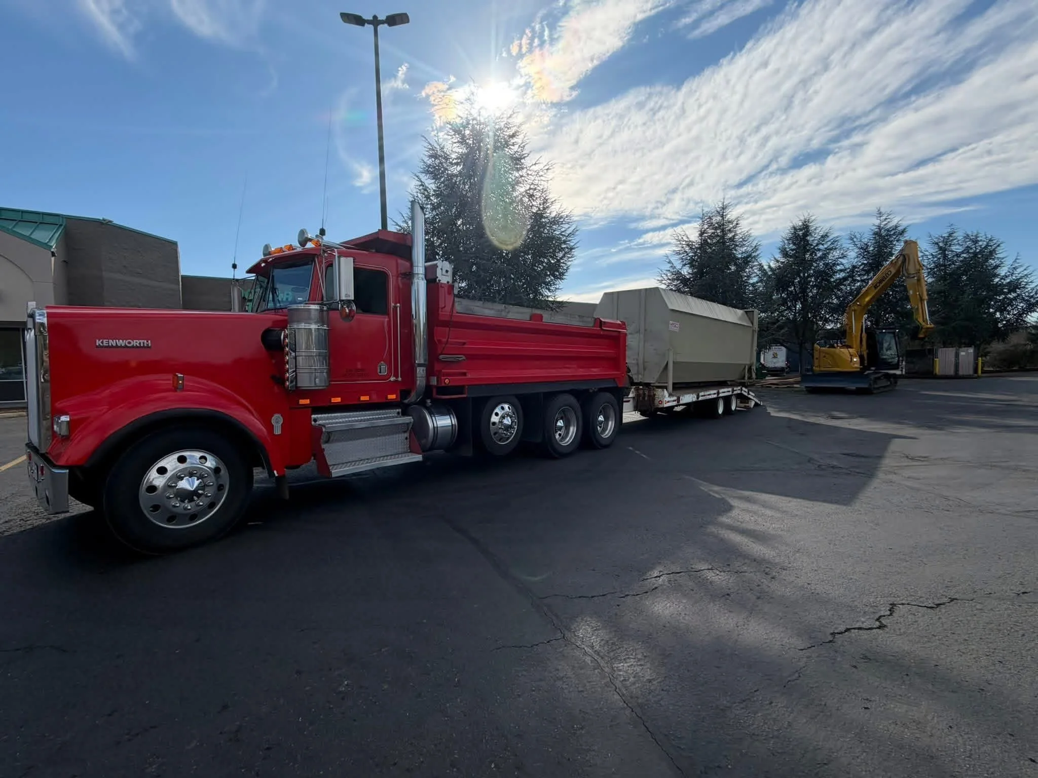 Red pickup truck with a trailer carrying a large container parked in a parking lot, with a yellow excavator in the background and trees under a partly cloudy sky.