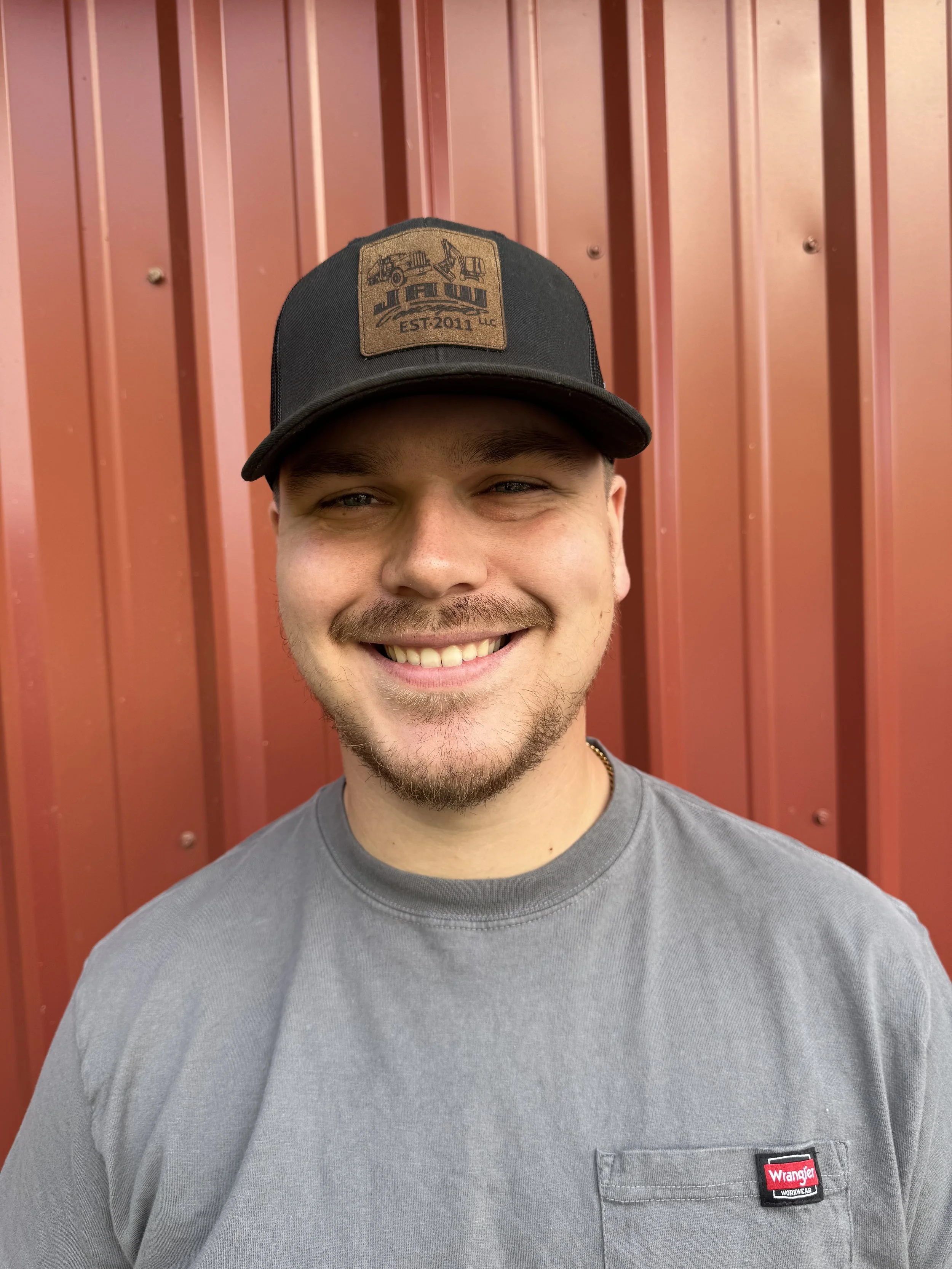 A young man with a beard and mustache smiling, wearing a black baseball cap with a logo, and a gray Wrangler t-shirt, standing in front of a red metal wall.