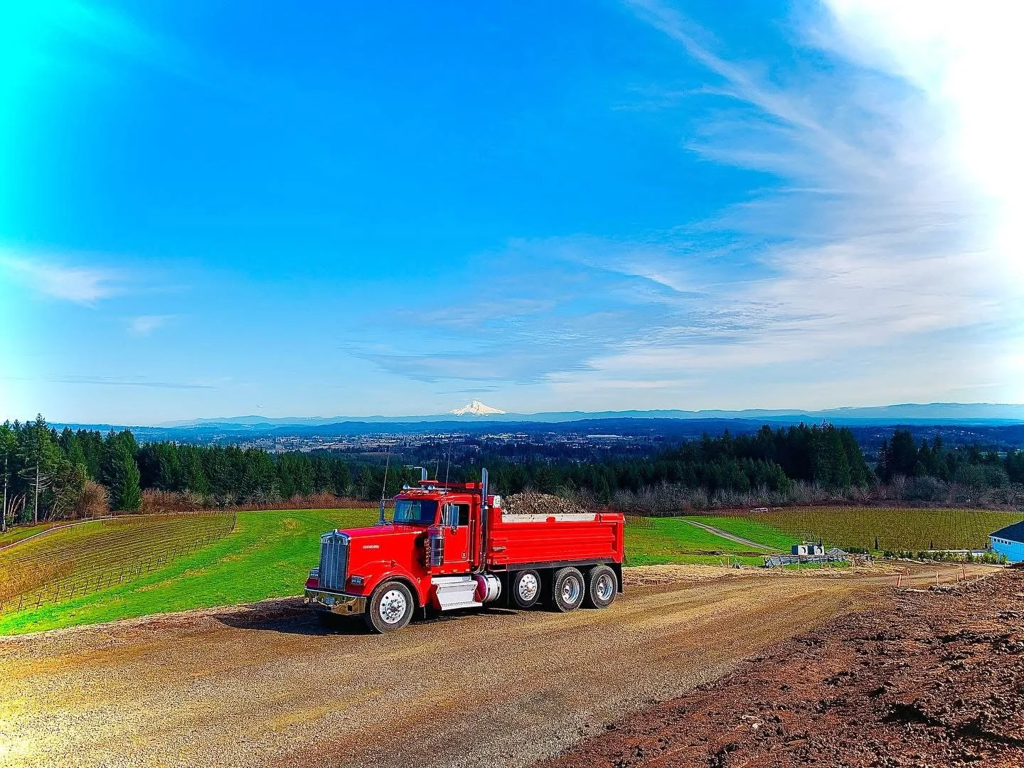 A red truck parked on a dirt road with green fields and trees in the background, a blue sky with clouds, and a distant snow-capped mountain.