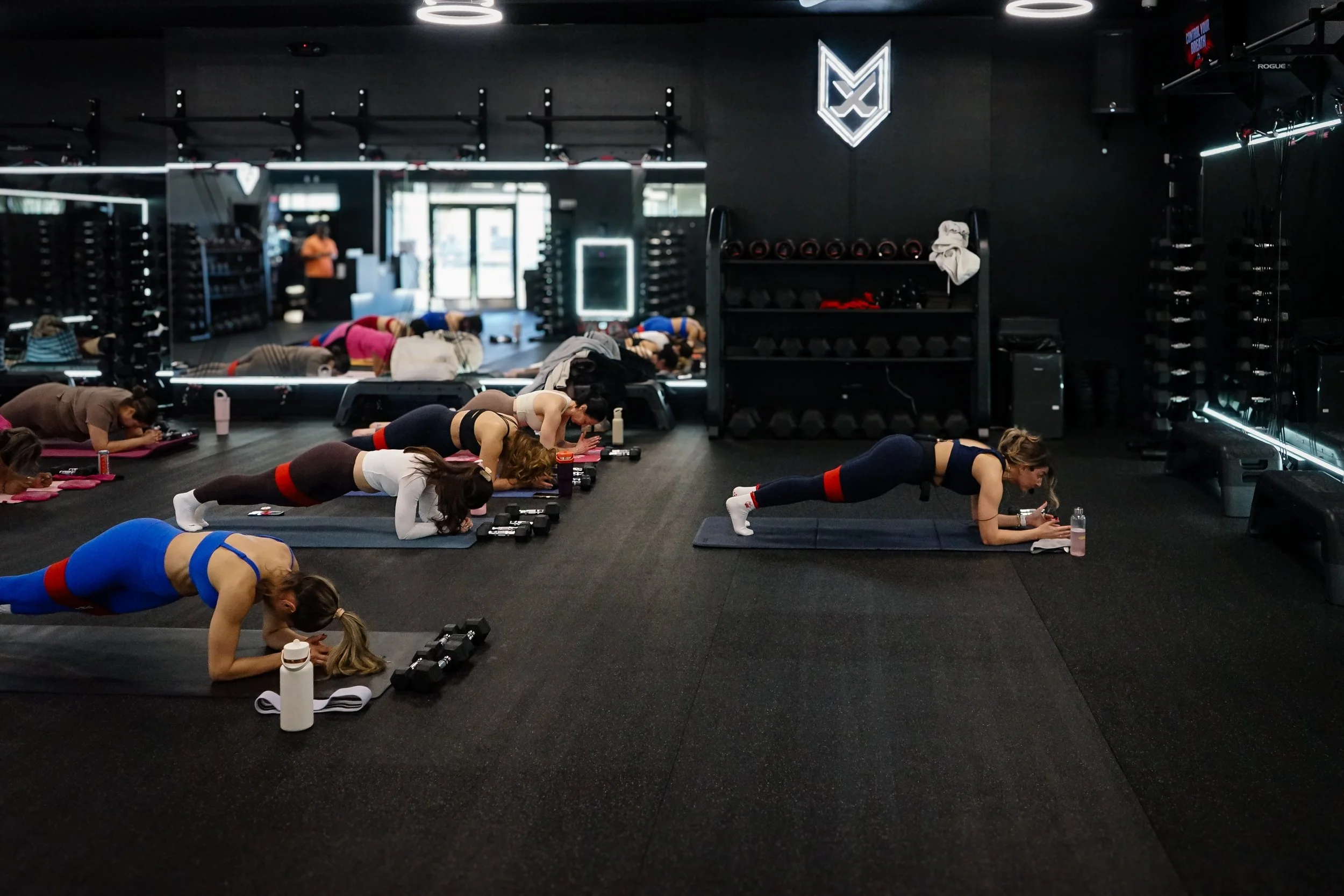 People participating in a group fitness class doing planks on yoga mats in a dark gym with black walls, mirrors, and fitness equipment.