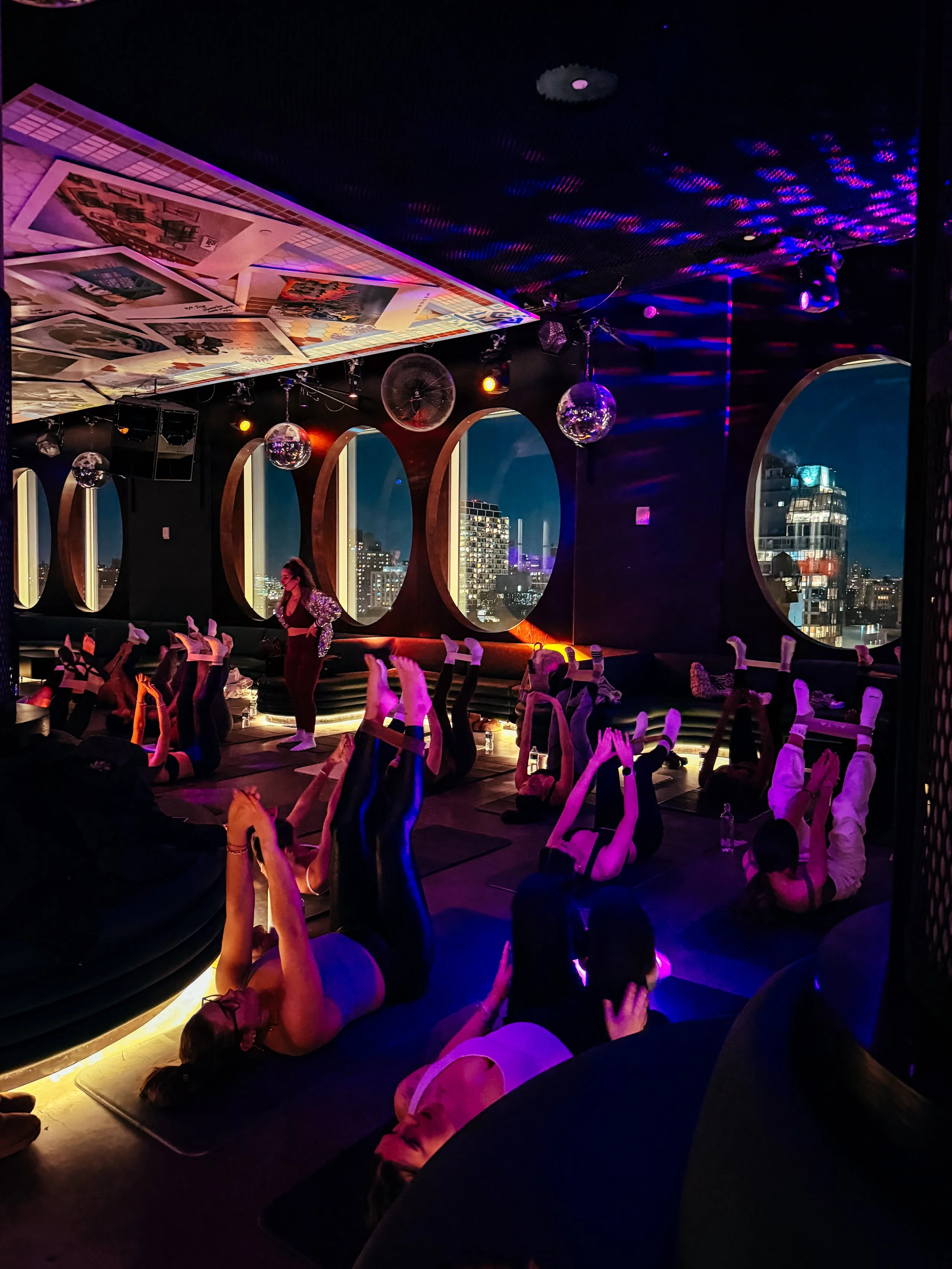 People participating in a yoga or exercise class in a dimly lit room with city views through large windows, colorful lights, and disco balls.