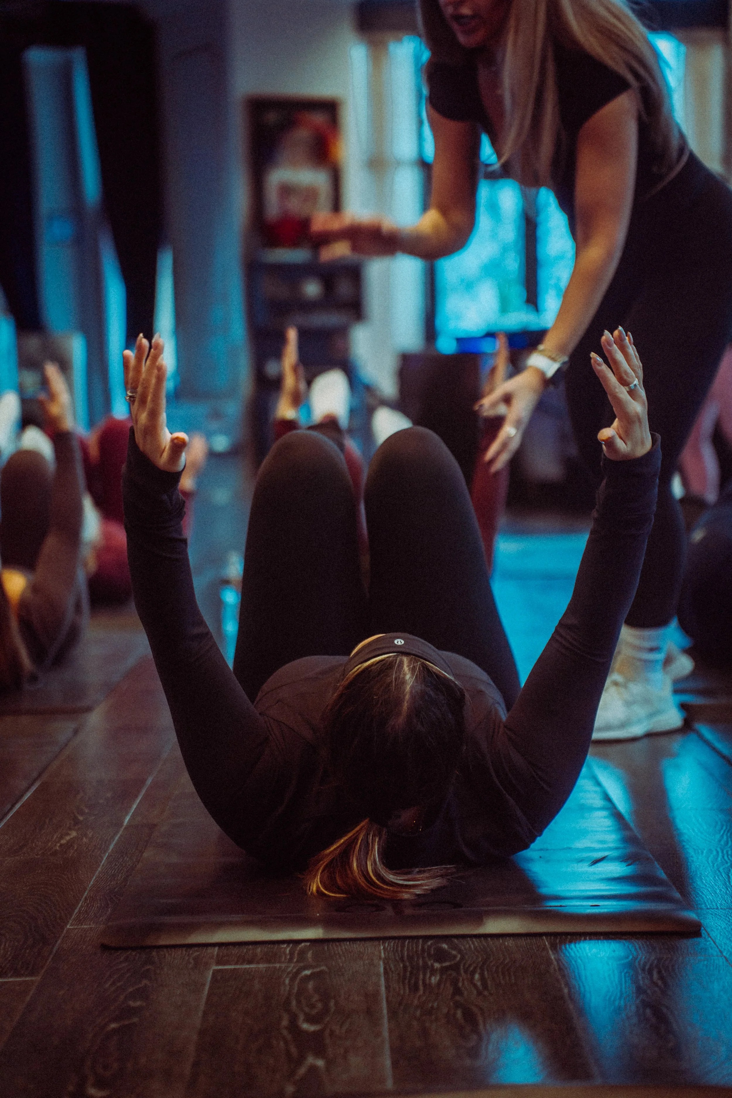 Person lying on yoga mat doing a reverse plank pose, with arms raised and head and shoulders elevated, in a room with wooden flooring and people sitting on mats in the background.