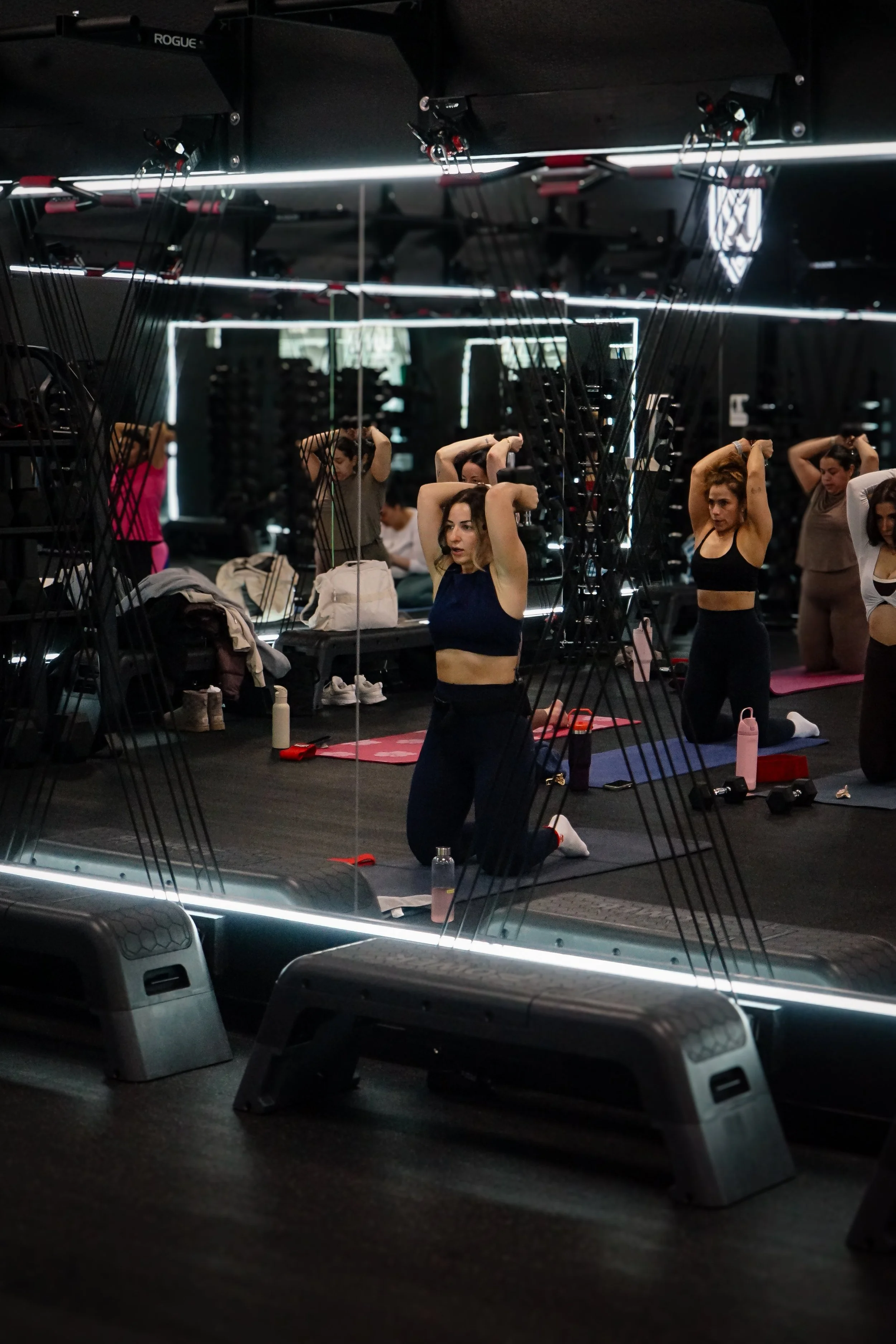 Women participating in a fitness class doing stretches on yoga mats in a dimly lit gym with mirrors and fitness equipment.