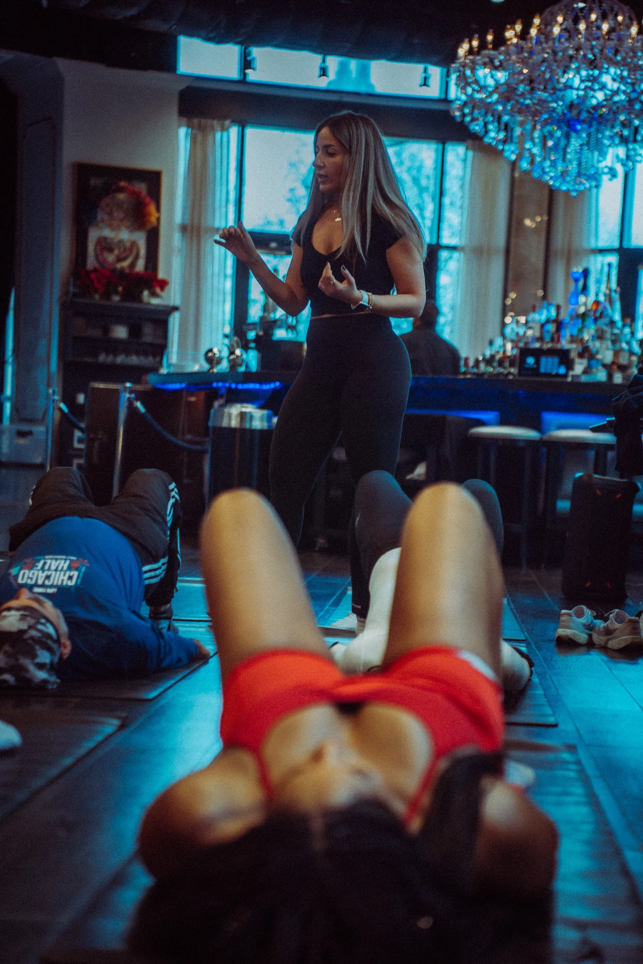 A woman standing and speaking in a dimly lit room with a chandelier, large windows, and various decorations, while people lie on the floor in front of her. The photo is taken from the perspective of someone lying down, with legs and shoes visible at 