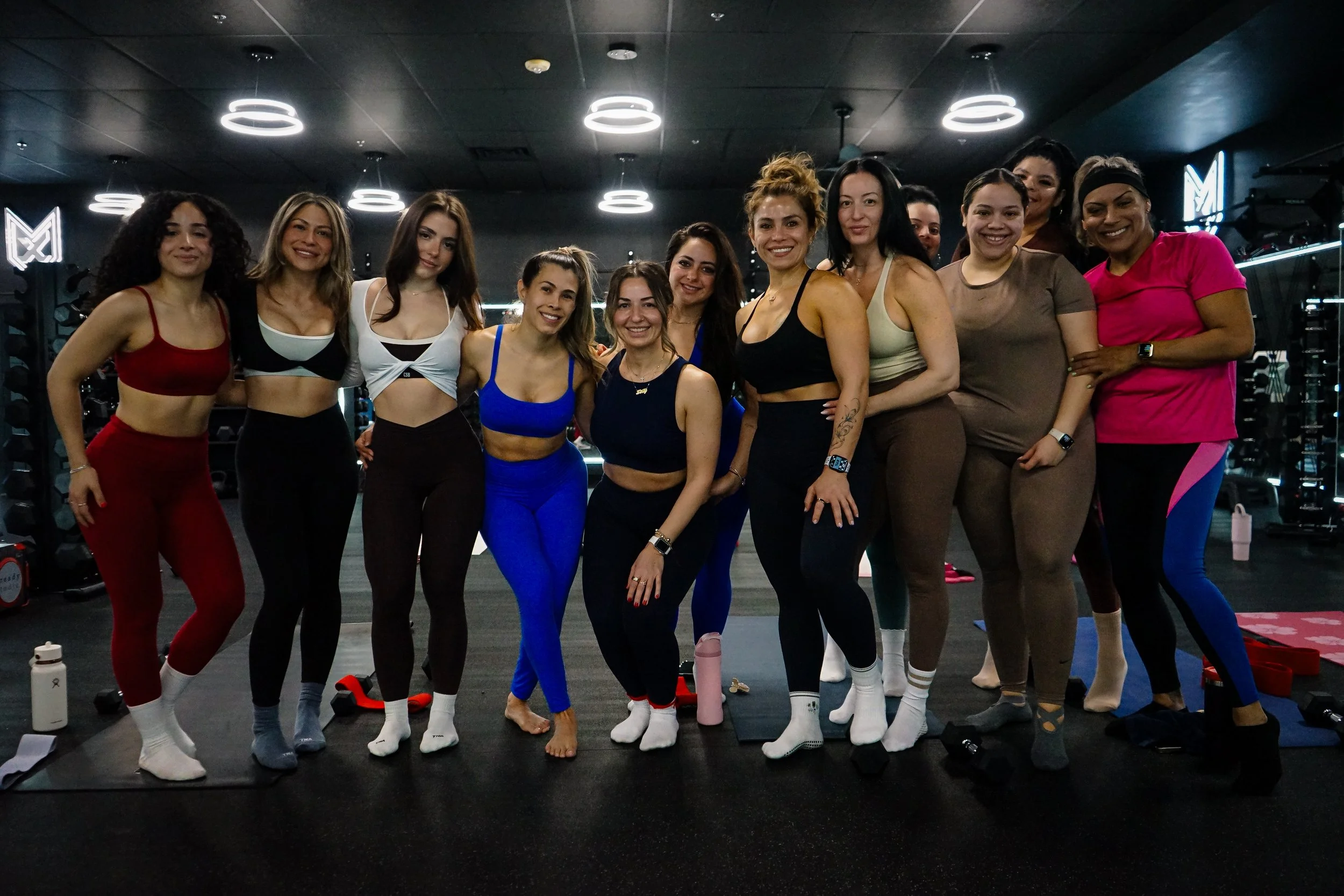 A group of women dressed in colorful workout clothes standing together in a fitness studio, smiling at the camera.