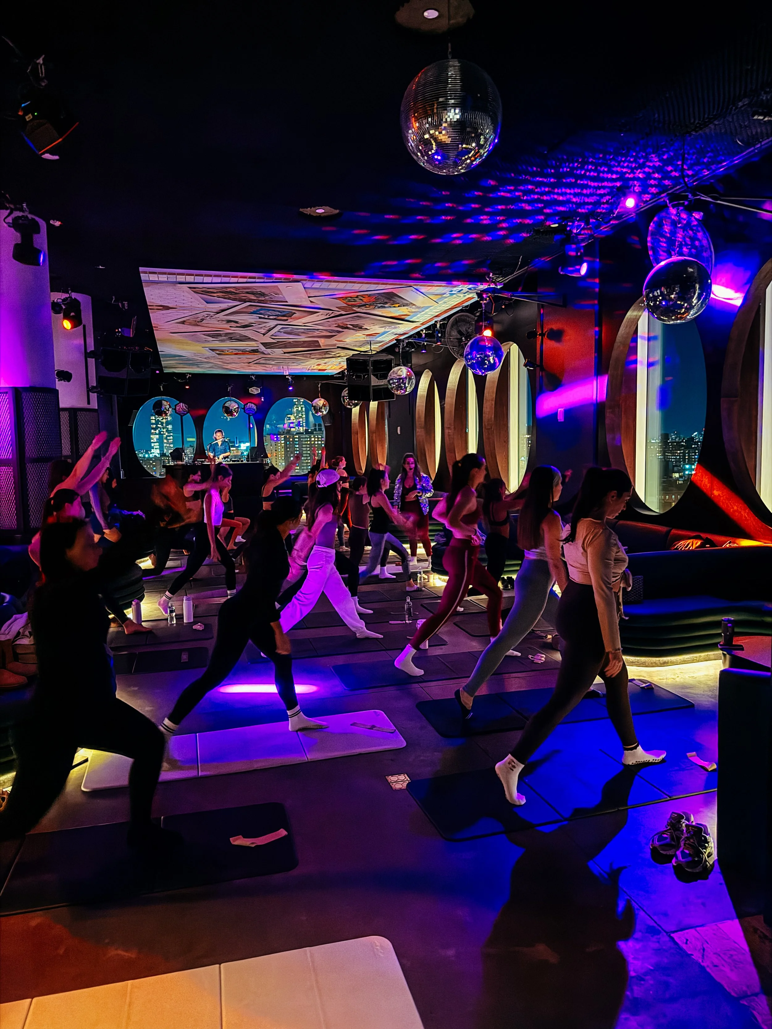 People participating in a yoga class in a dark, colorful studio with a city skyline view through large windows, featuring a disco ball and vibrant lighting.