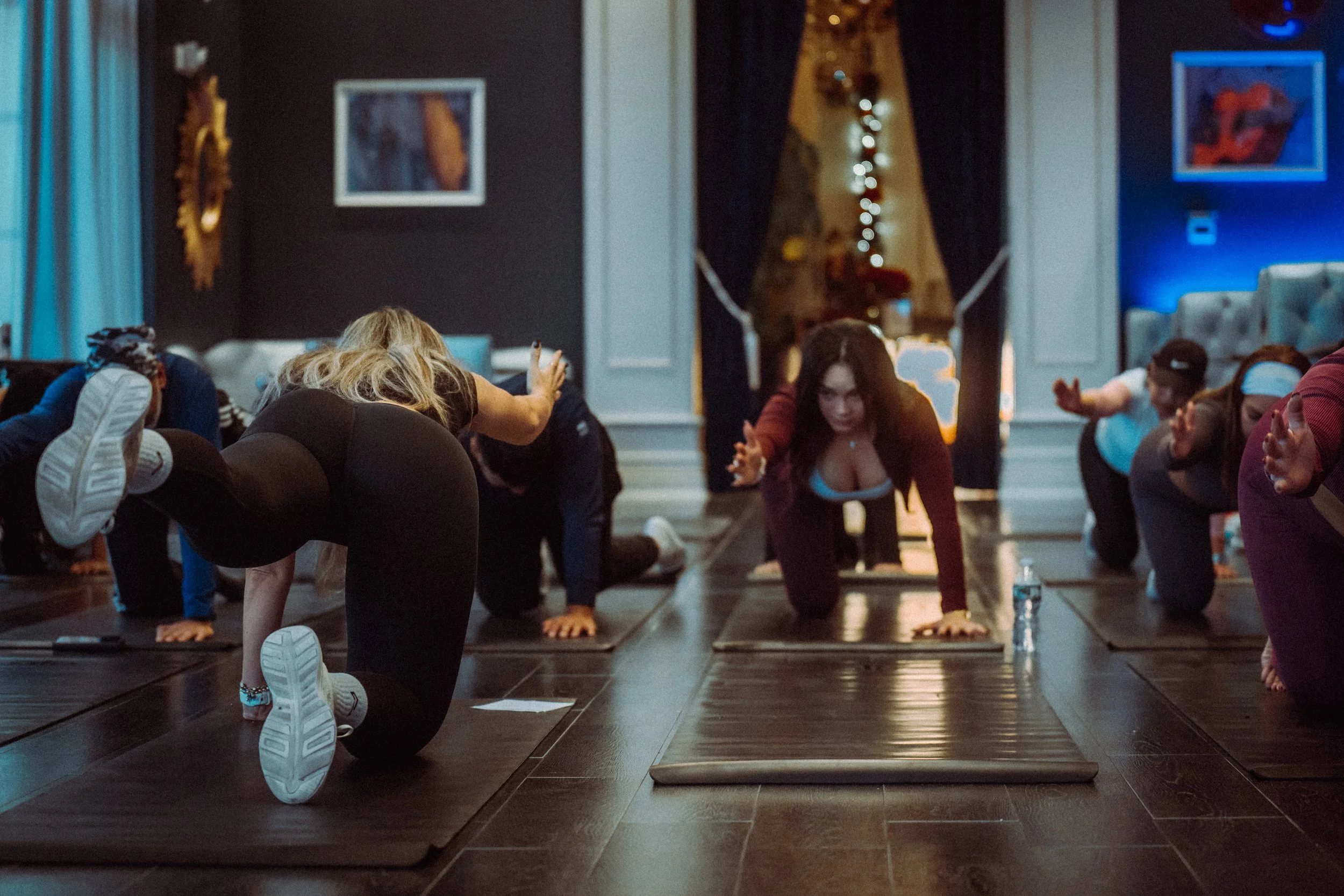 People participating in a yoga or fitness class, doing a pose on mats in a dimly lit room with some decorative lighting and artwork on the walls.