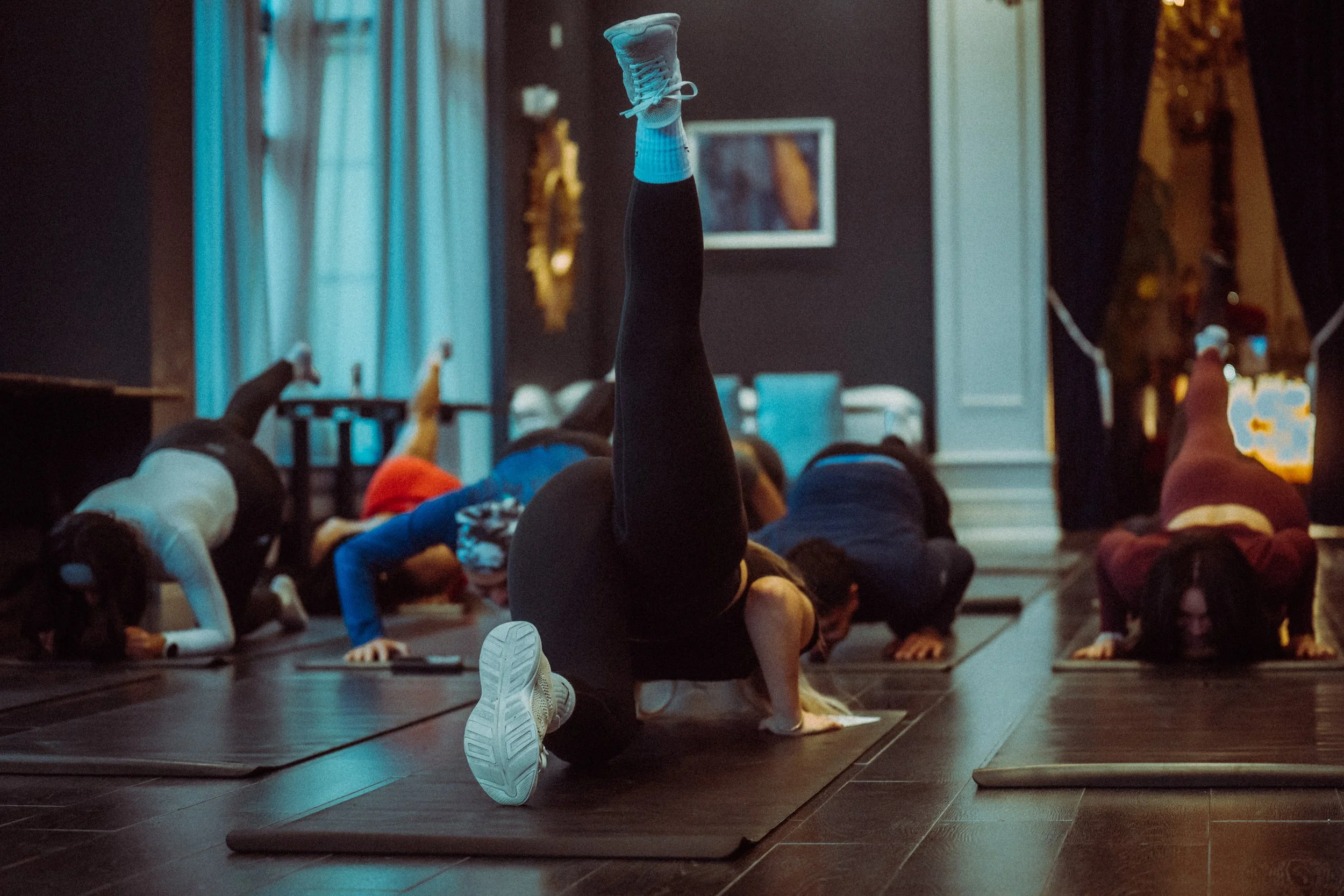 A group fitness class with participants lying on exercise mats, one person raising a leg during a workout, in a dimly lit room with artwork and a large mirror on the wall.