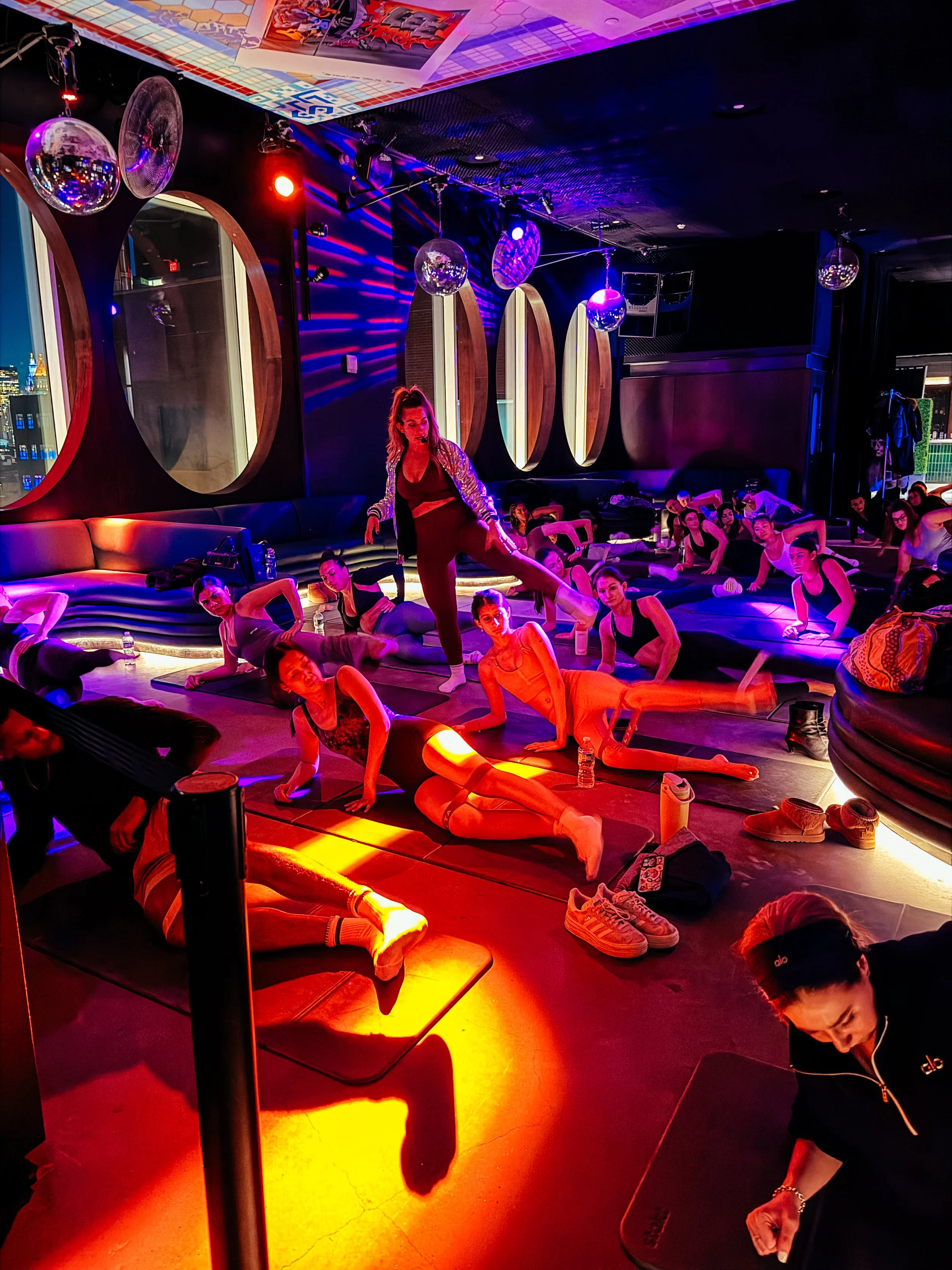 A group fitness class, likely yoga or stretching, taking place in a dimly lit studio with vibrant colored lighting, mirrors on the wall, and participants lying or sitting on mats while a woman instructor stands in the center.