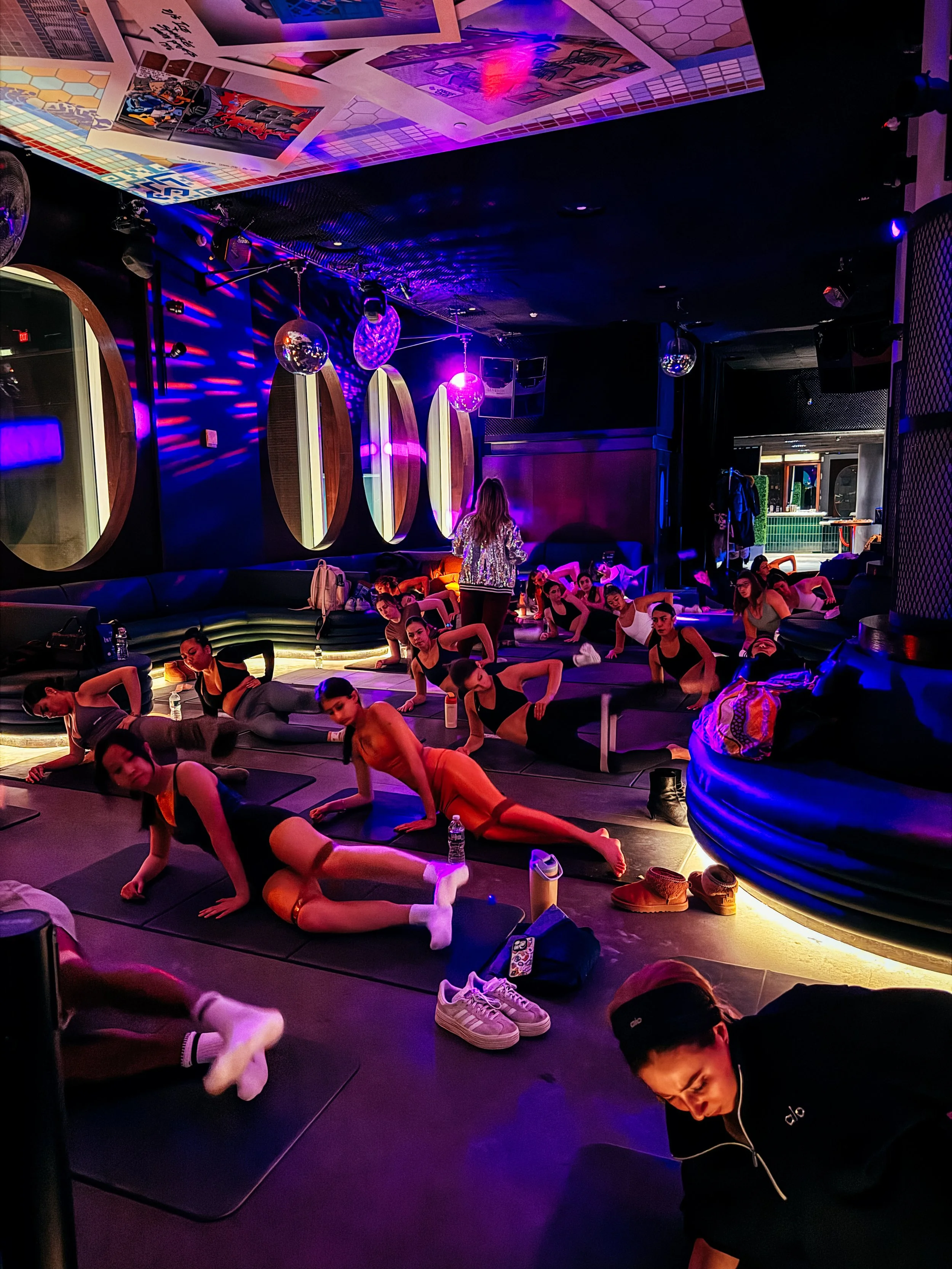 A group of people doing yoga or stretching exercises on mats in a dimly lit room with colorful lighting and disco balls, suggesting a dance or fitness studio.