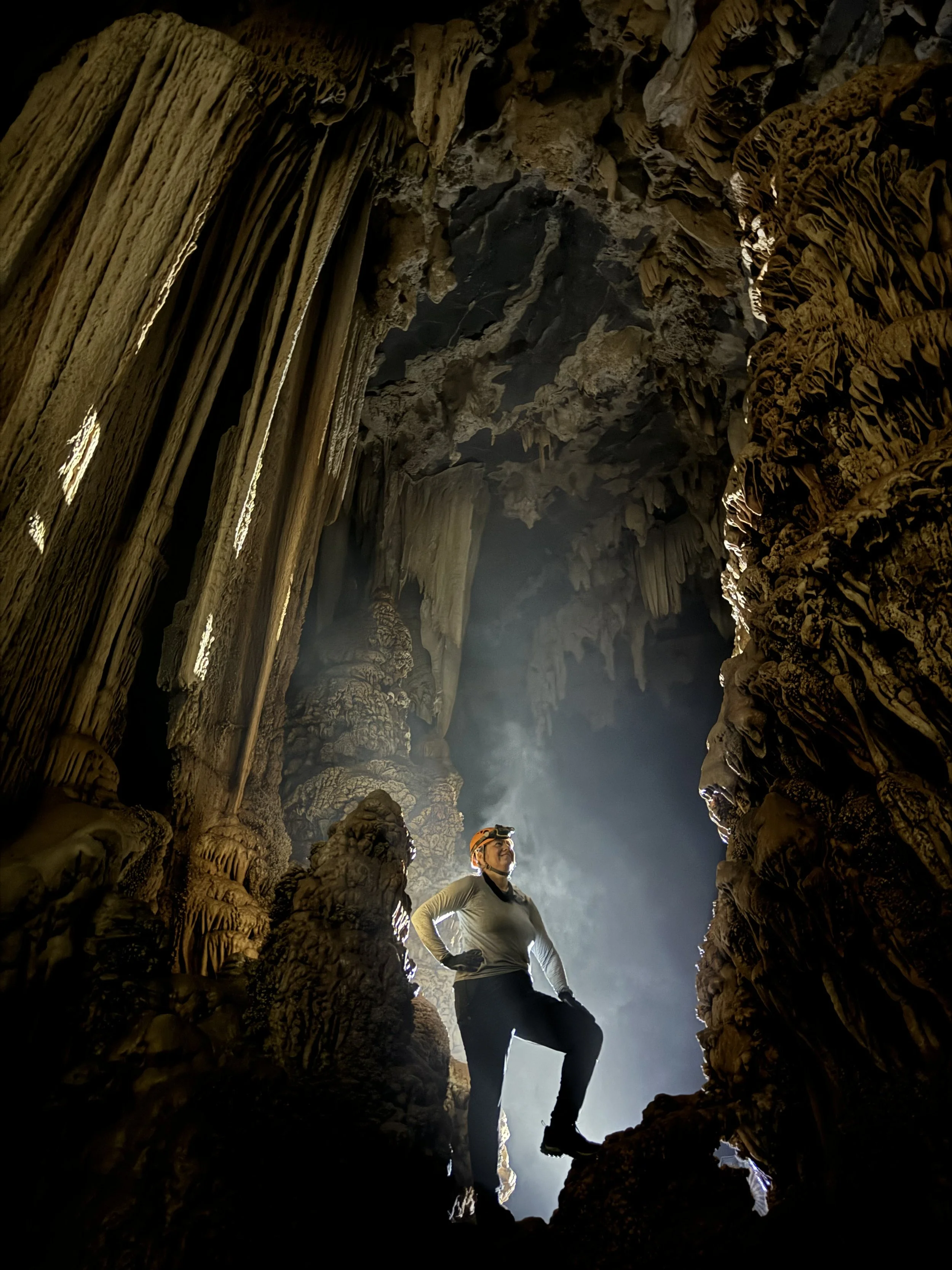 Amanda Mortimer is standing inside a large cave with stalactites and stalagmites, wearing a headlamp and gear, illuminated by natural light.