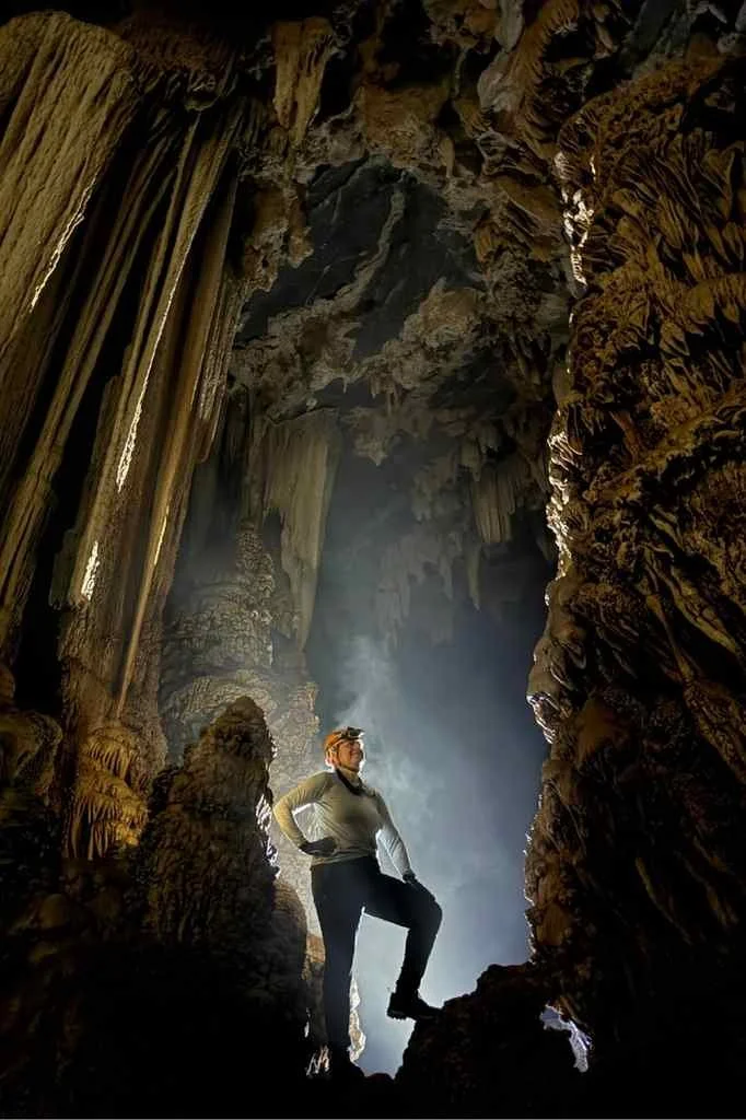 Amanda Mortimer is standing inside a large cave with stalactites and stalagmites, wearing a headlamp and gear, illuminated by natural light.