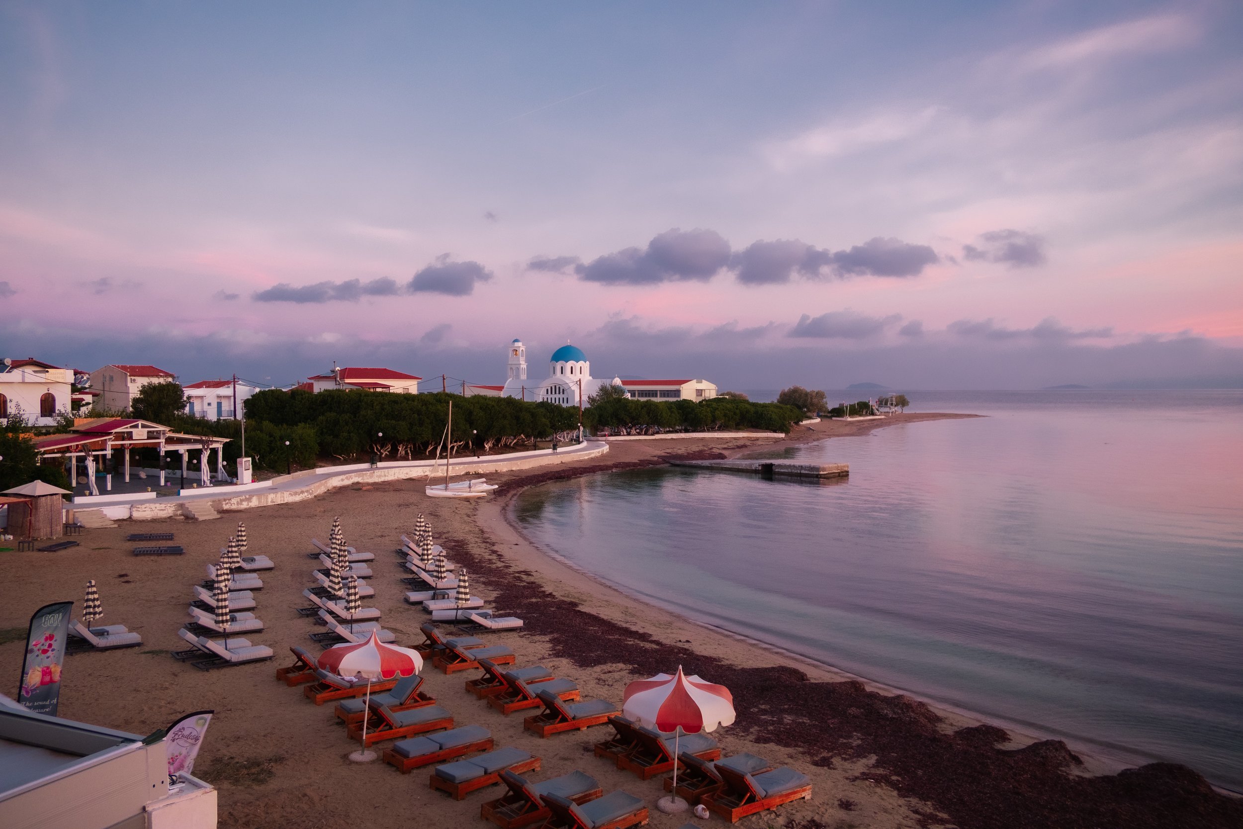 Greek seaside village beach at sunset with striped umbrellas, lounge chairs, white buildings, and a blue-domed church