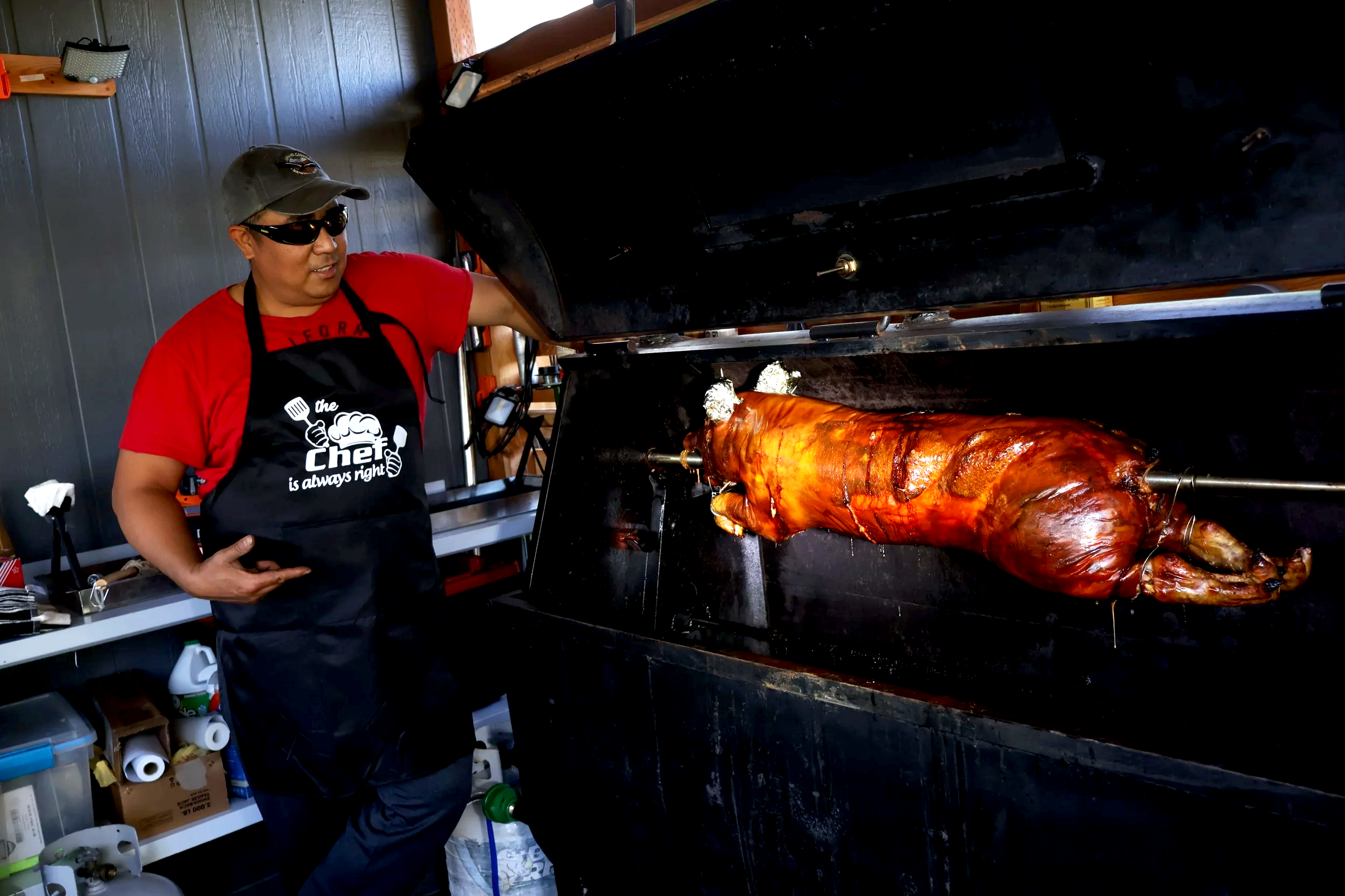 A man wearing a gray cap, sunglasses, a red T-shirt, and a black apron with the words 'the chef is always right' standing next to a large roasted pig on a spit inside a workshop or garage.
