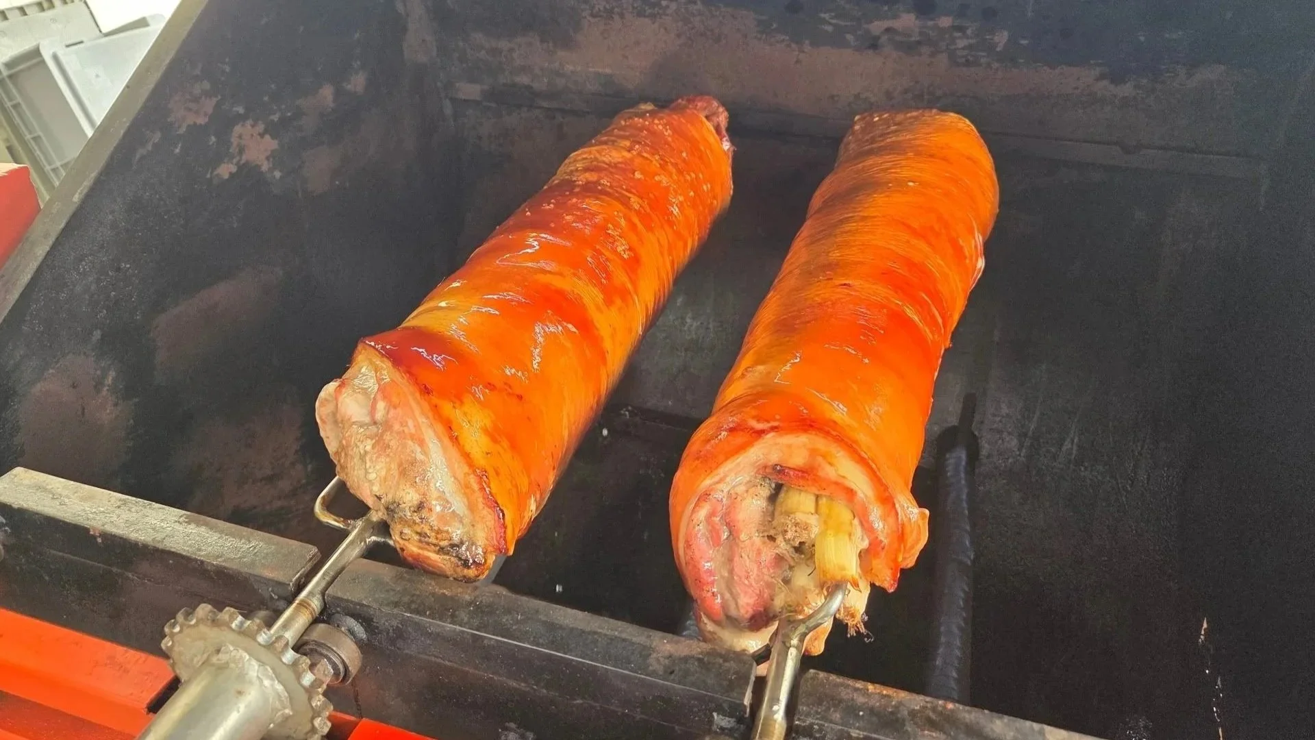 Two large pig roasts wrapped in orange pork skin cooking on a rotisserie over a black grill or smoker.