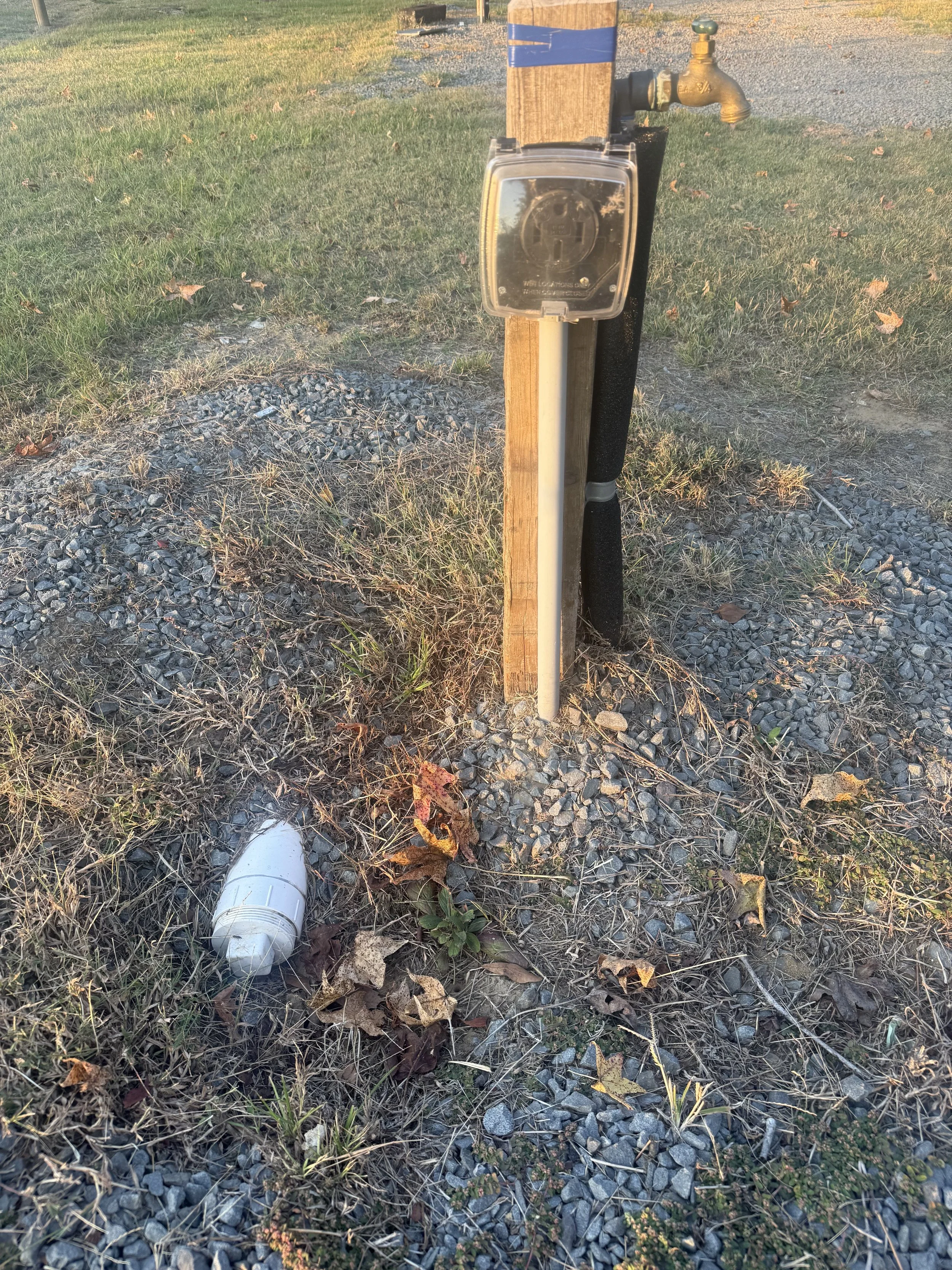 A wooden utility pole with an electric outlet box and a brass water faucet attached, set in a gravel and grassy area, with a fallen leaf in the foreground.