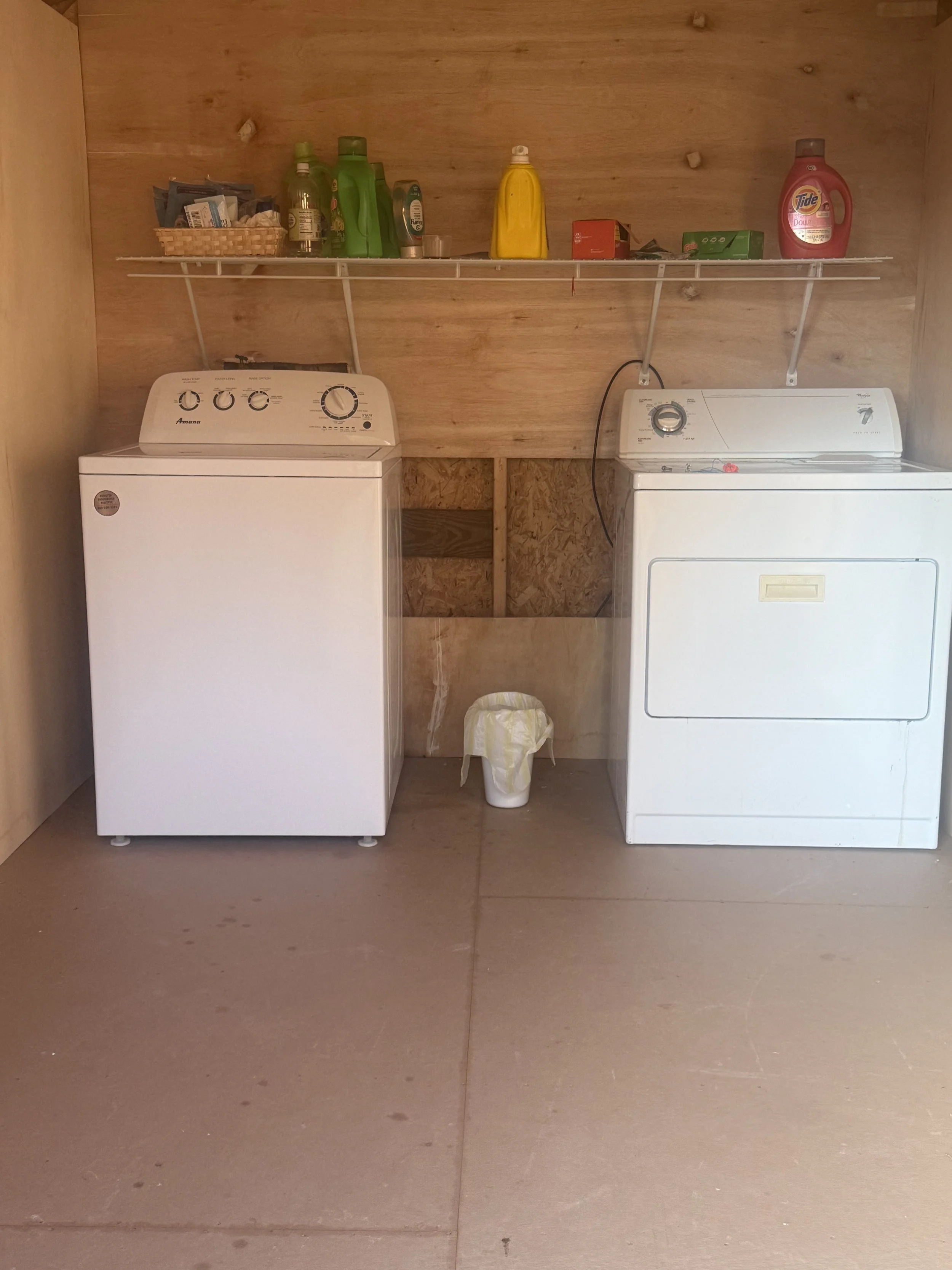 A laundry room with a washing machine on the left and a dryer on the right, both white, set against a wooden wall. A shelf above holds laundry supplies, including detergent bottles, a basket with small items, and other containers. A small trash can is positioned between the appliances.