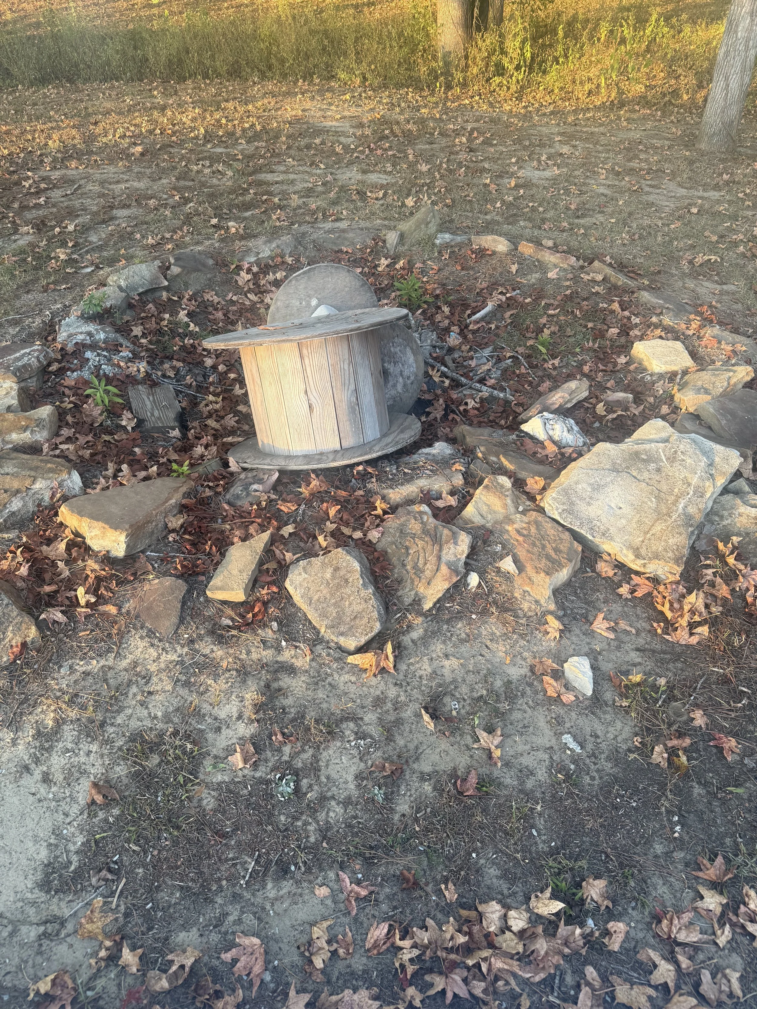 A small fire pit made of rocks with a wooden spool used as a table inside, surrounded by fallen autumn leaves, in an outdoor setting with trees and grass in the background.