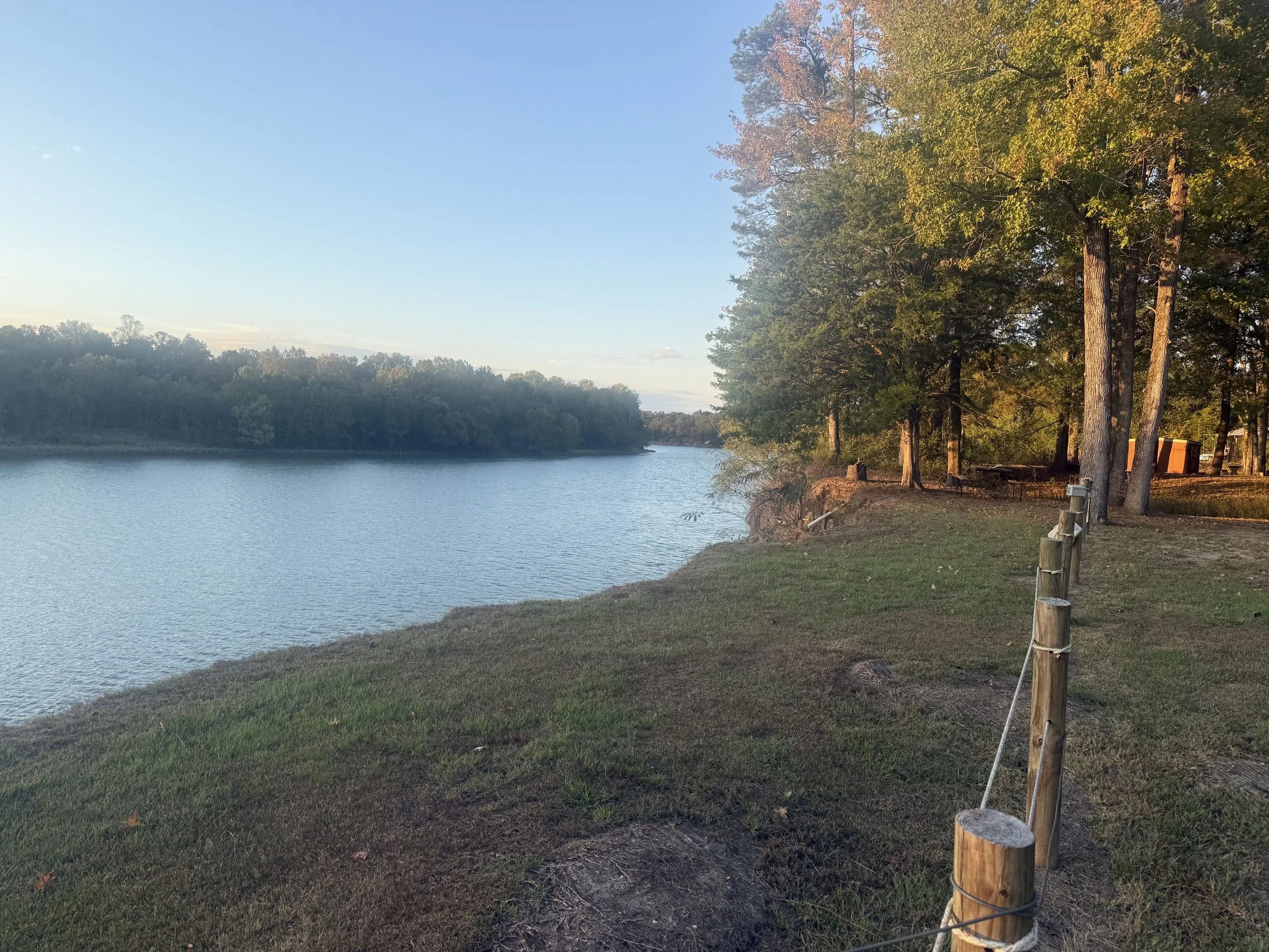 A lakeside scene during late afternoon with a calm body of water, green trees on the far shore, and a grassy area with a dirt path lined with wooden posts and a wire fence along the right side, with some trees casting shadows.