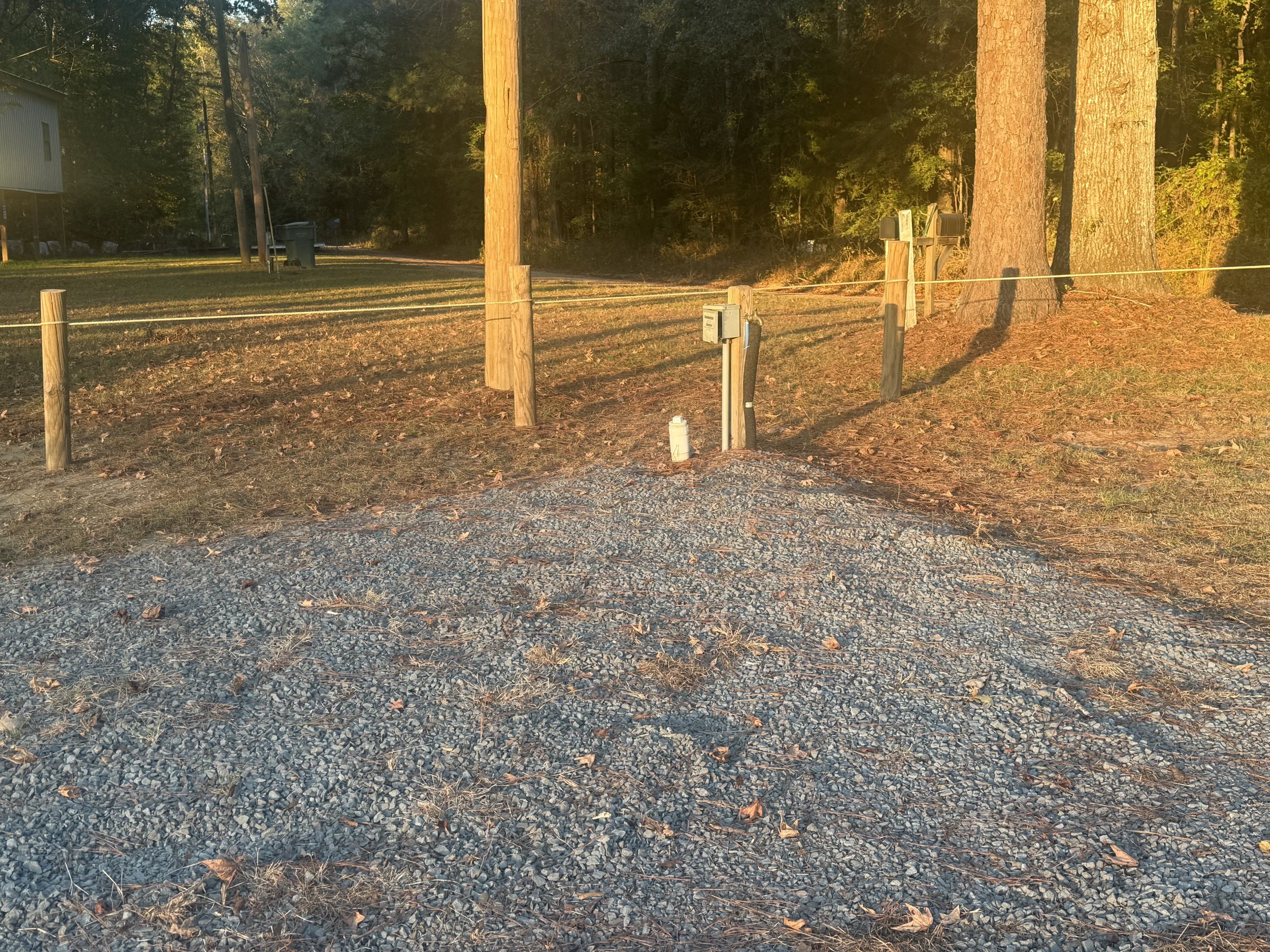 A gravel driveway with a white security light and a small white pipe near a wooden fence, with tall trees in the background, during sunset.