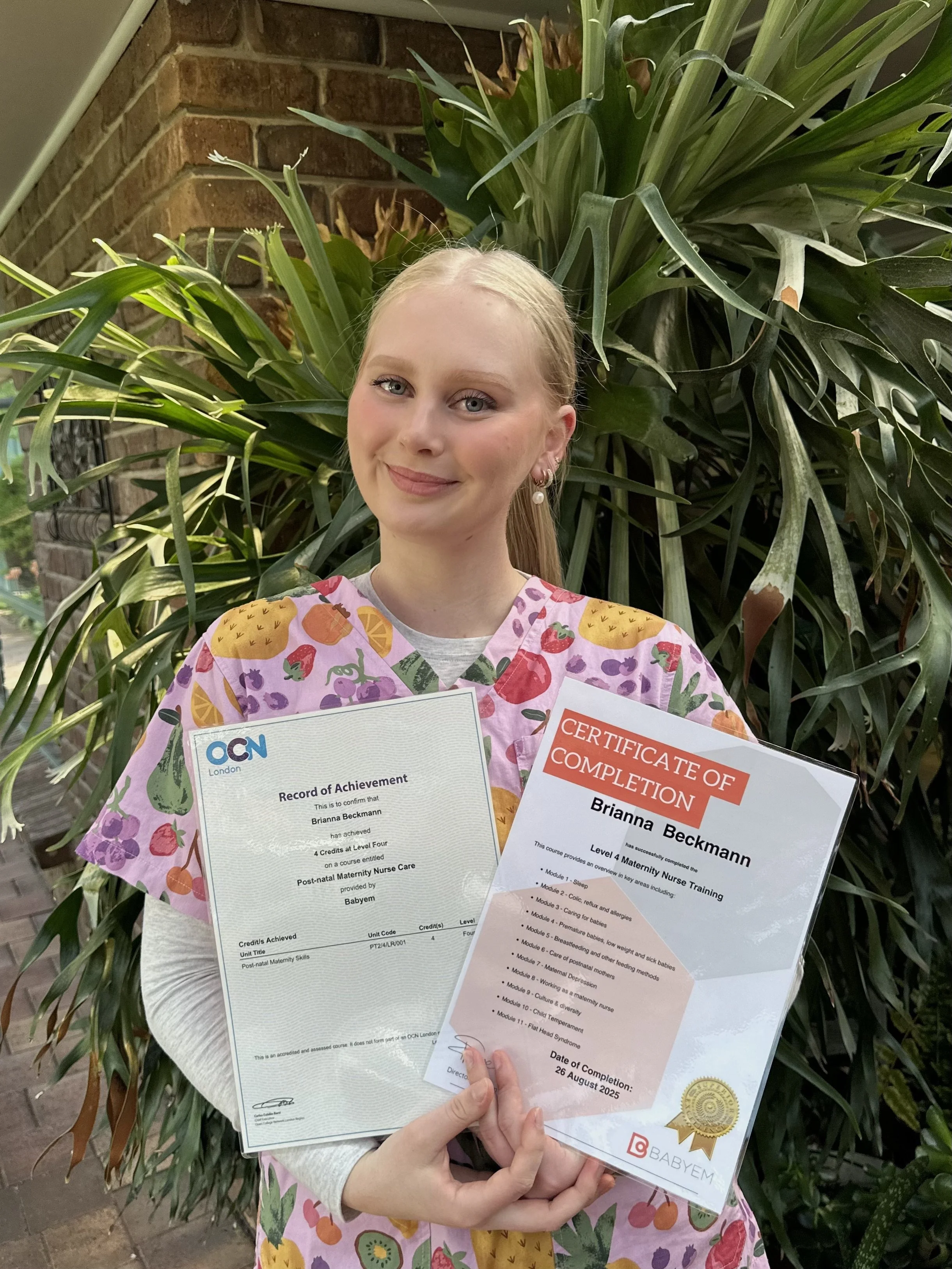 A young woman in colorful floral scrubs holding two certificates, standing in front of lush green plants and a brick wall, smiling at the camera.