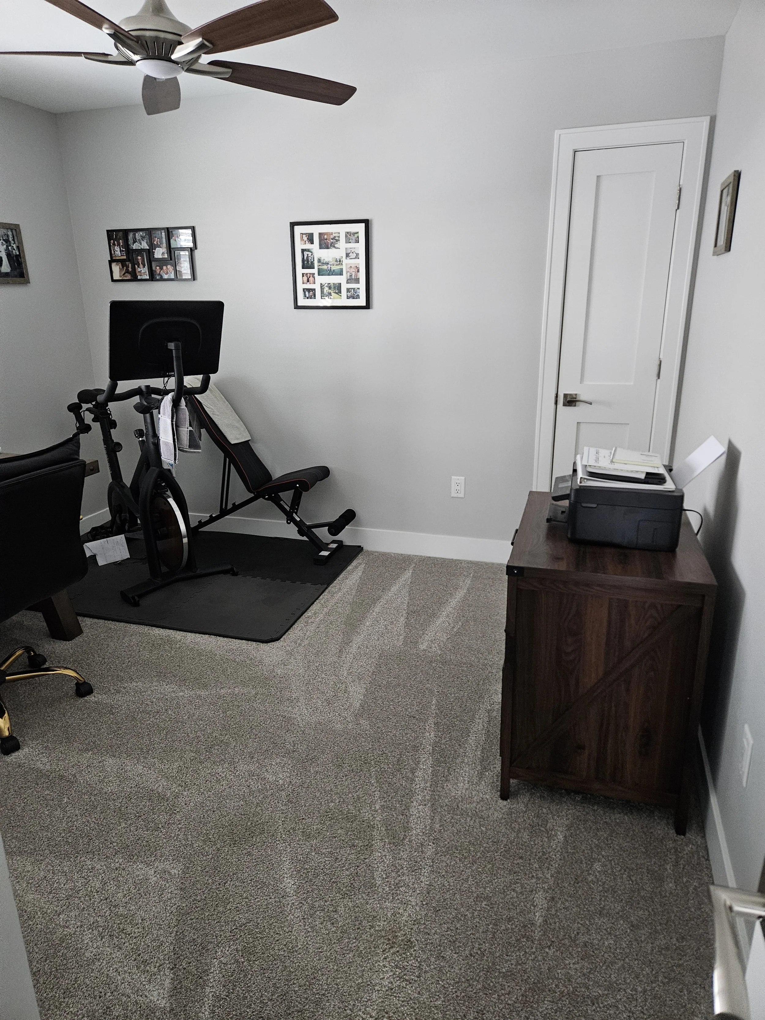 An office room with an exercise bike, a chair, a small wooden desk with a printer on top, and framed photos on the wall.