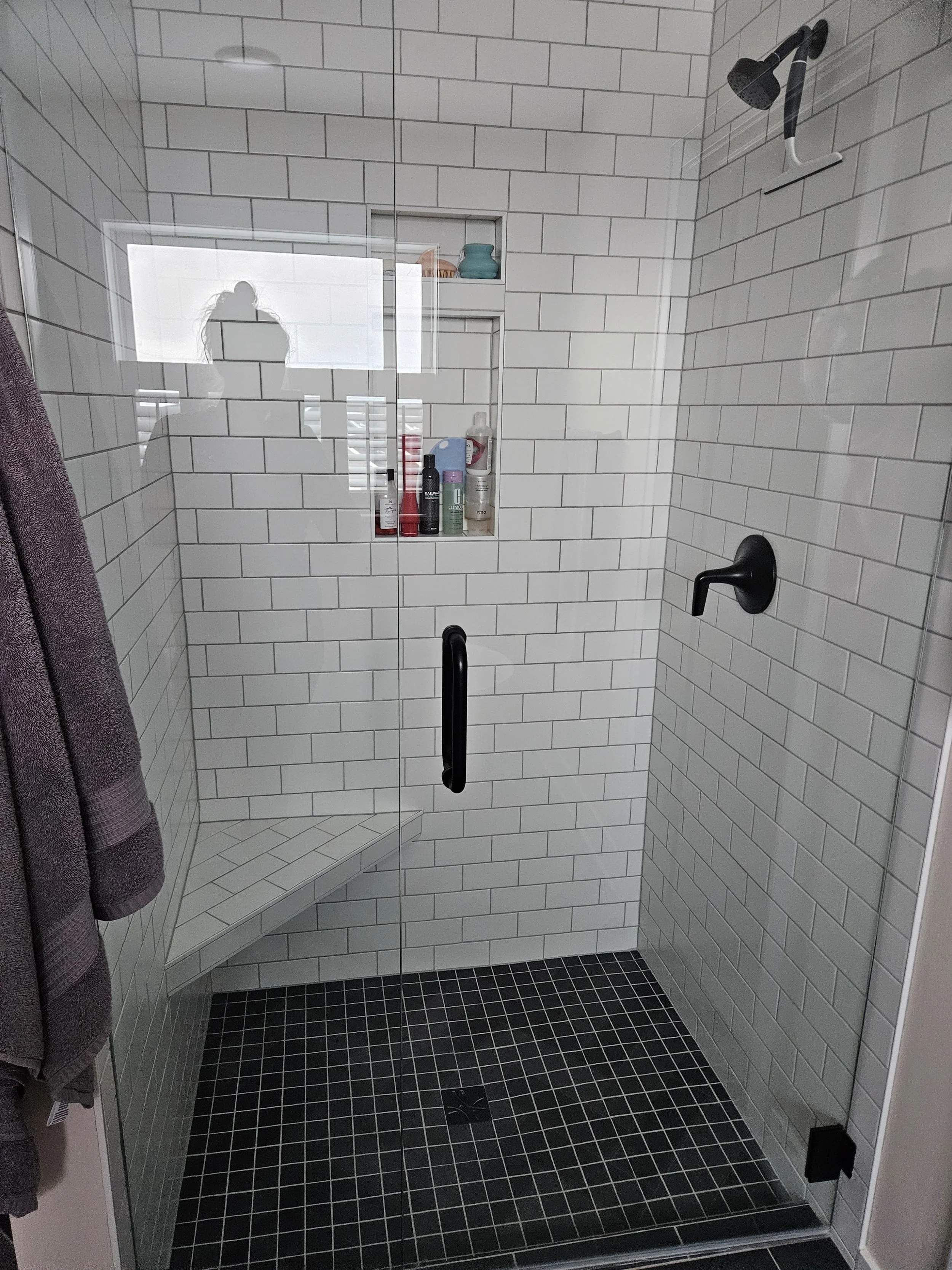 A modern walk-in shower with white subway tile walls, black fixtures, a built-in tiled bench, and a niche with bath products; a glass door with a black handle, and a window reflecting a silhouette of a person taking the photo.