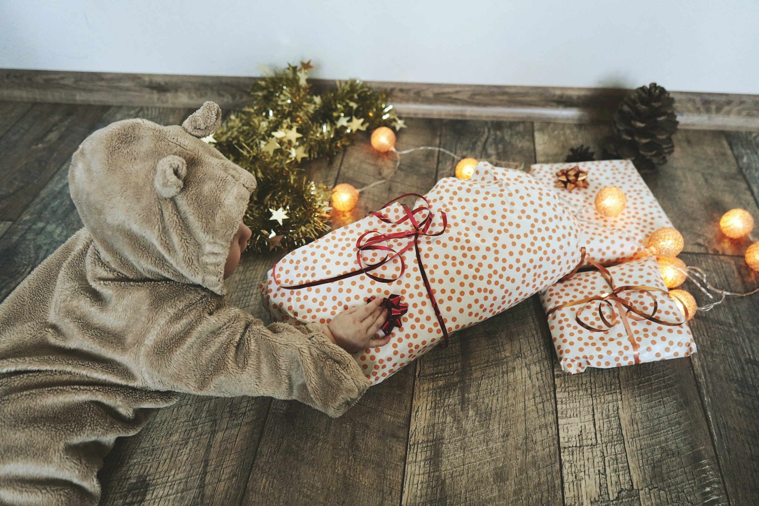 Child dressed in a bear costume reaching for wrapped Christmas presents on a wooden floor, with a small decorated Christmas tree and string lights in the background.