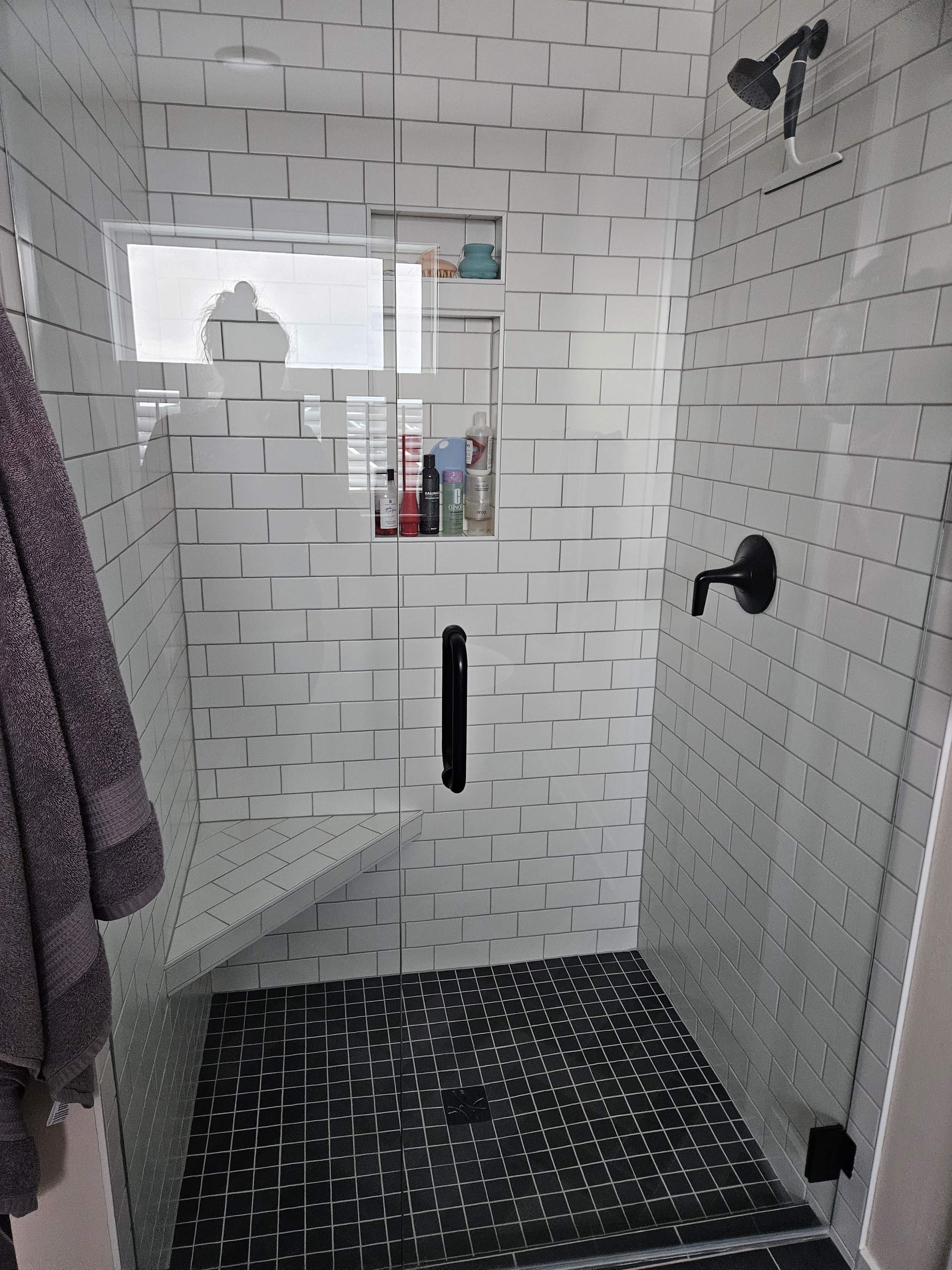 Modern walk-in shower with white subway tile walls, black fixtures, a glass door, a built-in corner niche with toiletries, and a black tiled floor.
