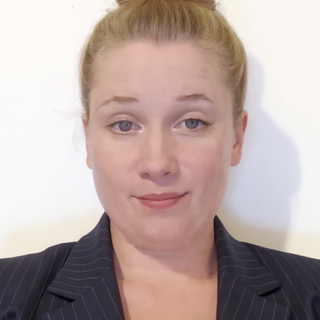 Close-up of a young woman with red hair and fair skin, wearing a dark pinstripe blazer, against a plain white background.