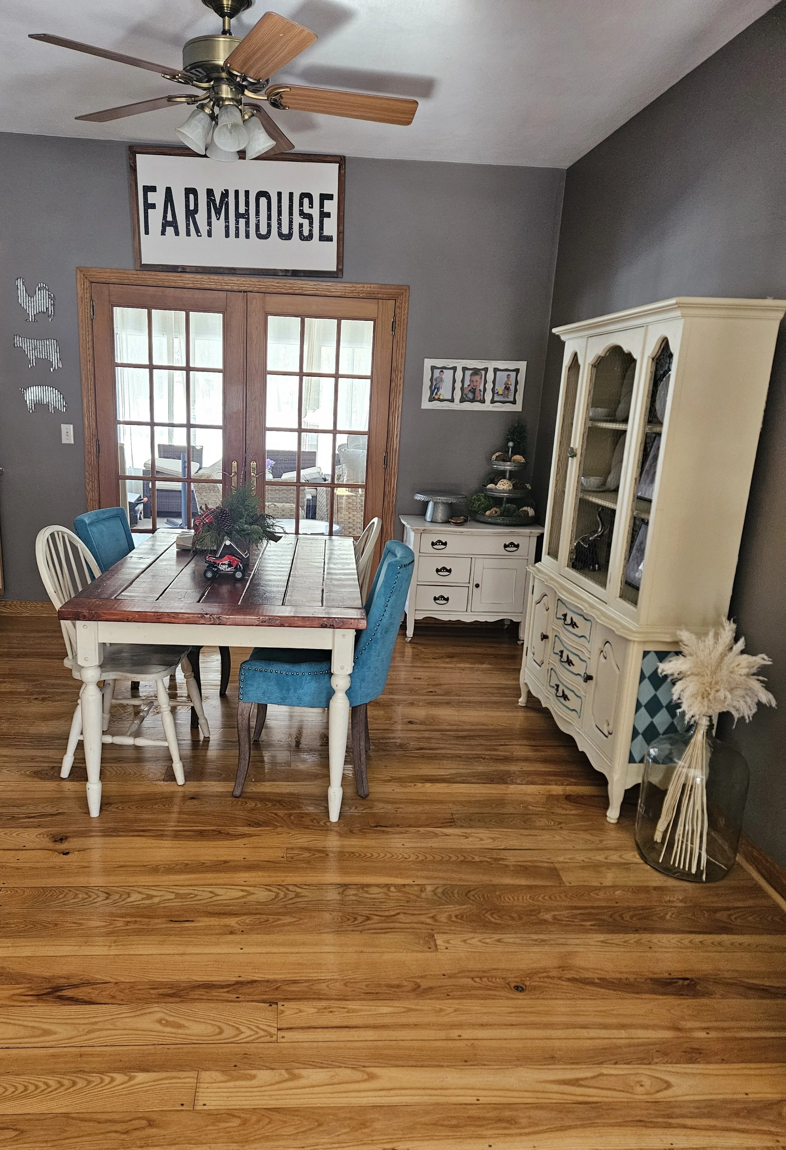 A dining room with gray walls, hardwood floor, and wooden French doors. There is a wooden dining table with mixed chairs, some with blue upholstery. A white china cabinet and a small white sideboard are in the room. Decor includes a large "FARMHOUSE" sign above the doors, animal-shaped metal wall art, framed photos, and a large glass vase with pampas grass.