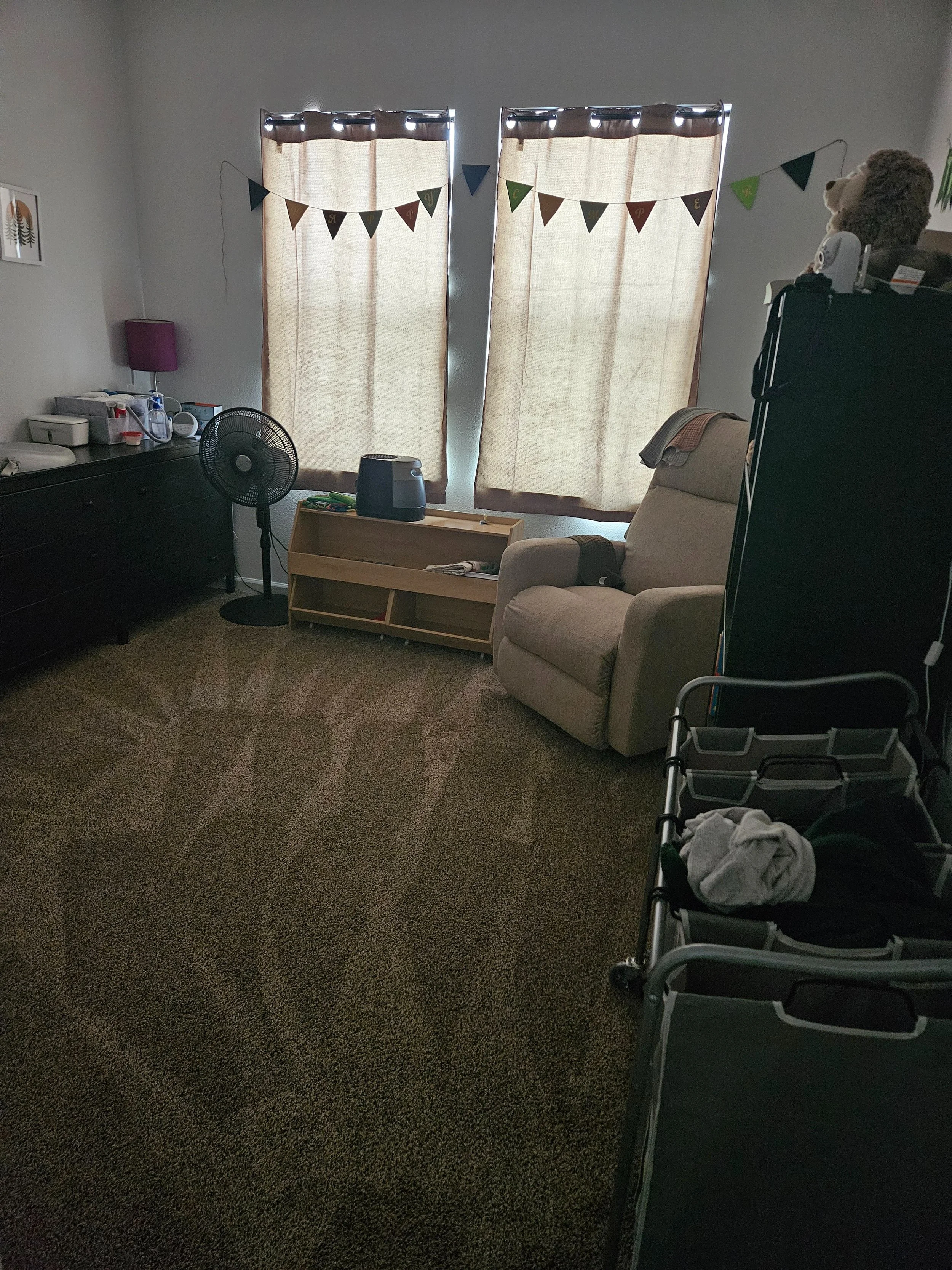 A living room with beige carpet, beige recliner, black dresser, standing fan, small bookshelf with items, and two windows with beige curtains. Decorations include a banner with colorful triangles and a stuffed bear on a black shelf.