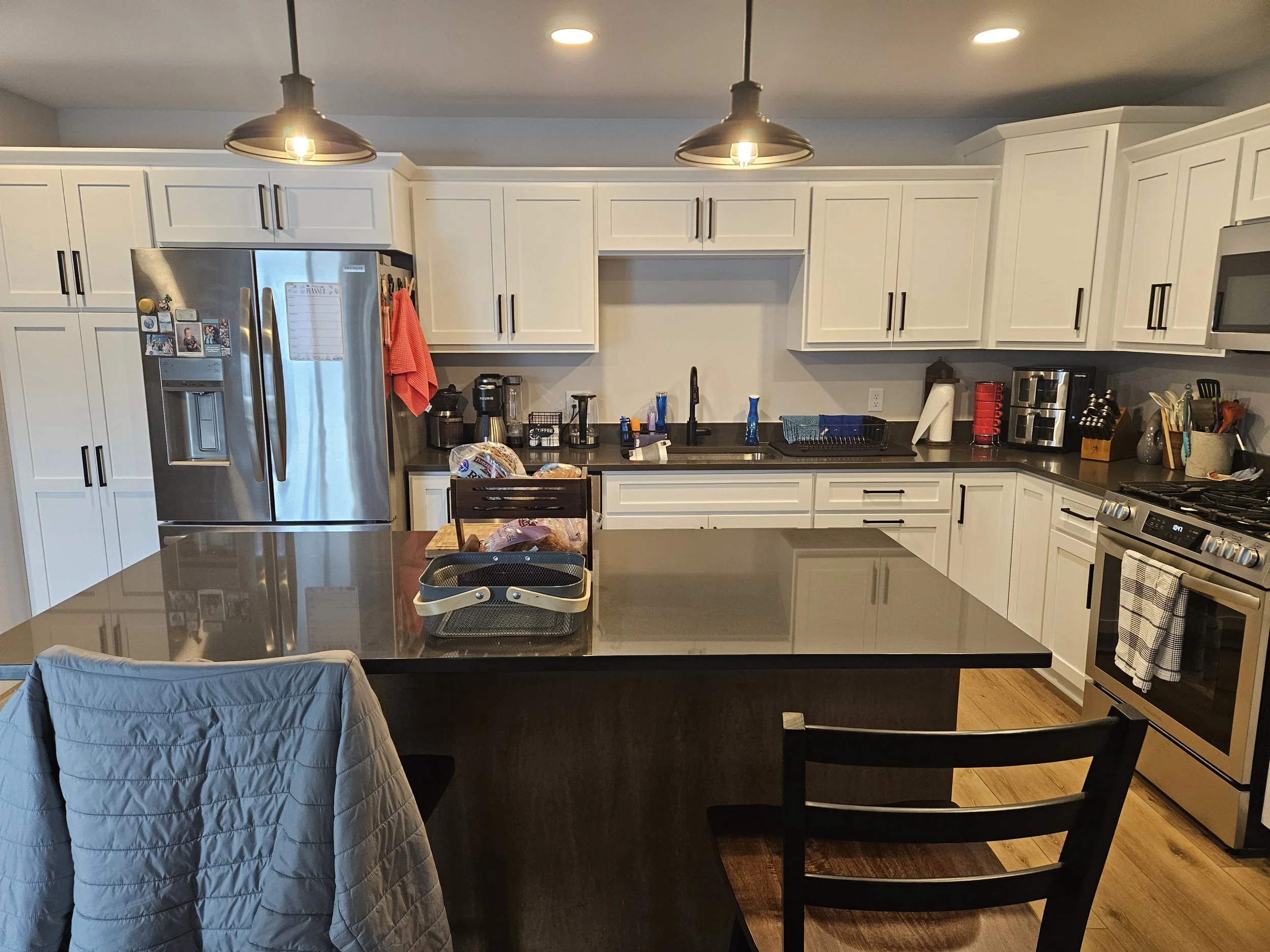 Modern kitchen with white cabinets, stainless steel refrigerator, black countertops, and various kitchen appliances. The island has a dark wood base and a gray countertop, with a beige jacket draped over a chair.